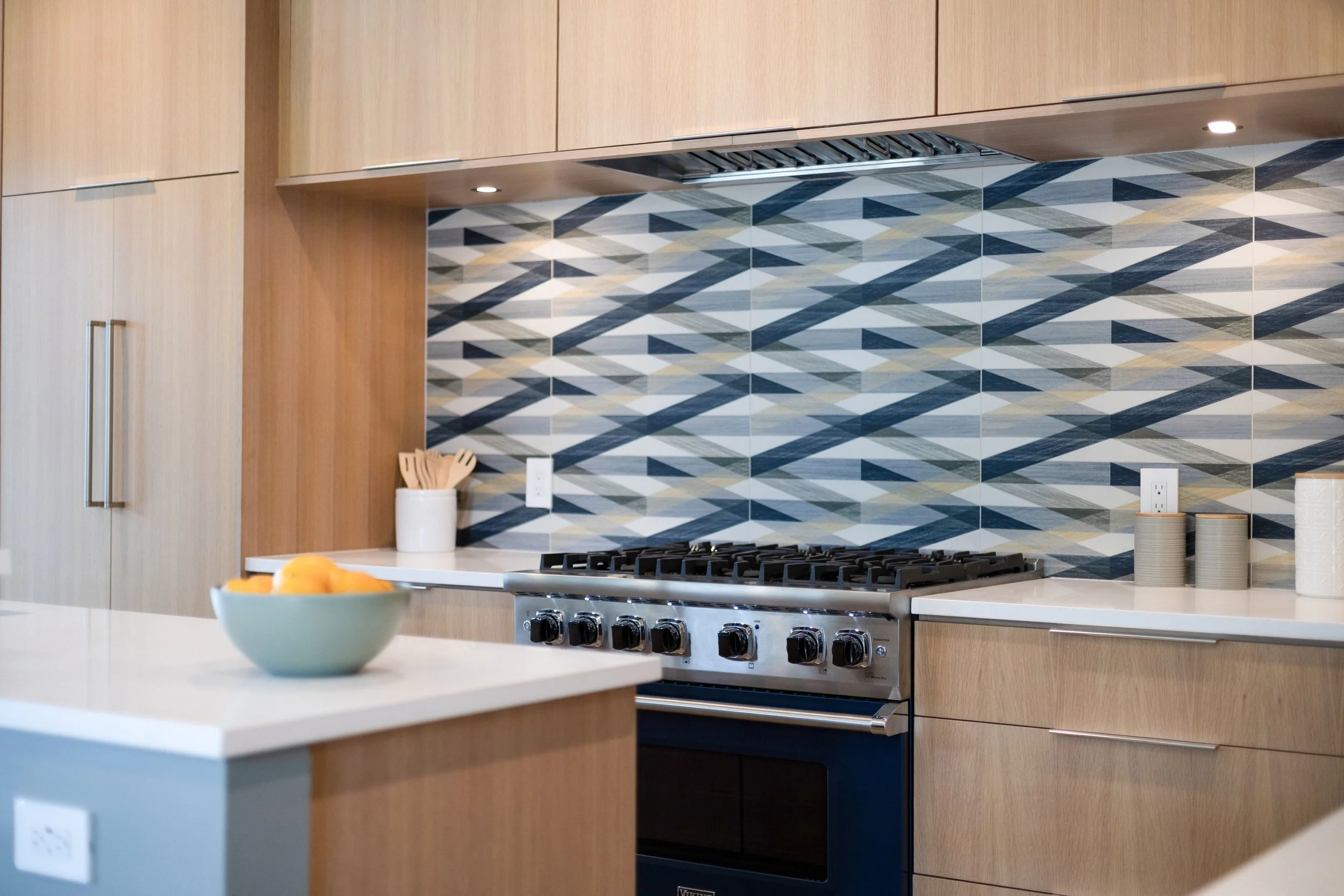 Modern kitchen with wooden cabinets, a geometric patterned tile backsplash in shades of blue, white, and gray, a stove, and countertop accessories like a container with wooden utensils and a bowl of fruit.
