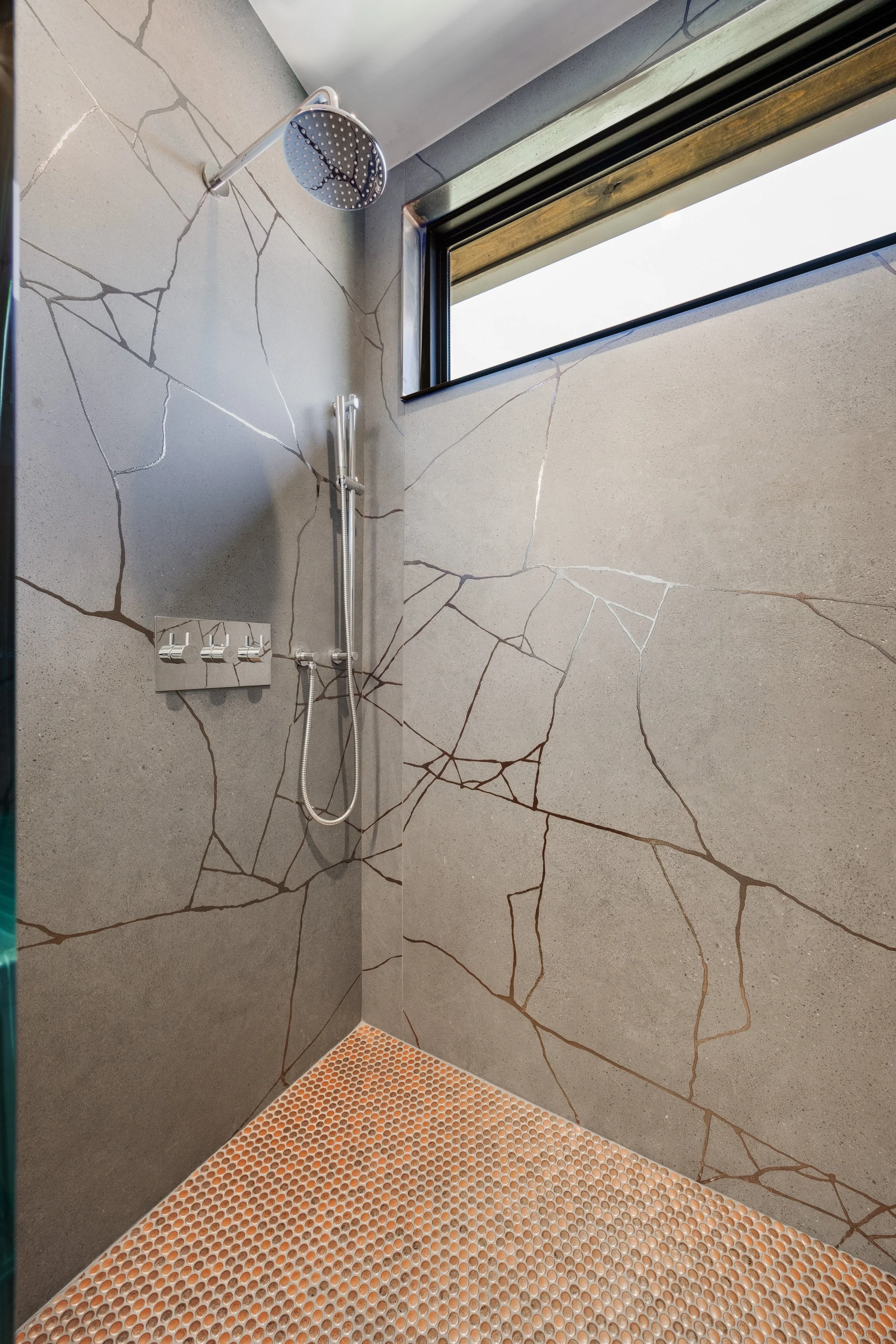 A shower with large gray stone-like tiles and a small window near the ceiling. There is a round showerhead, a hand-held shower nozzle, and three control knobs on the wall.