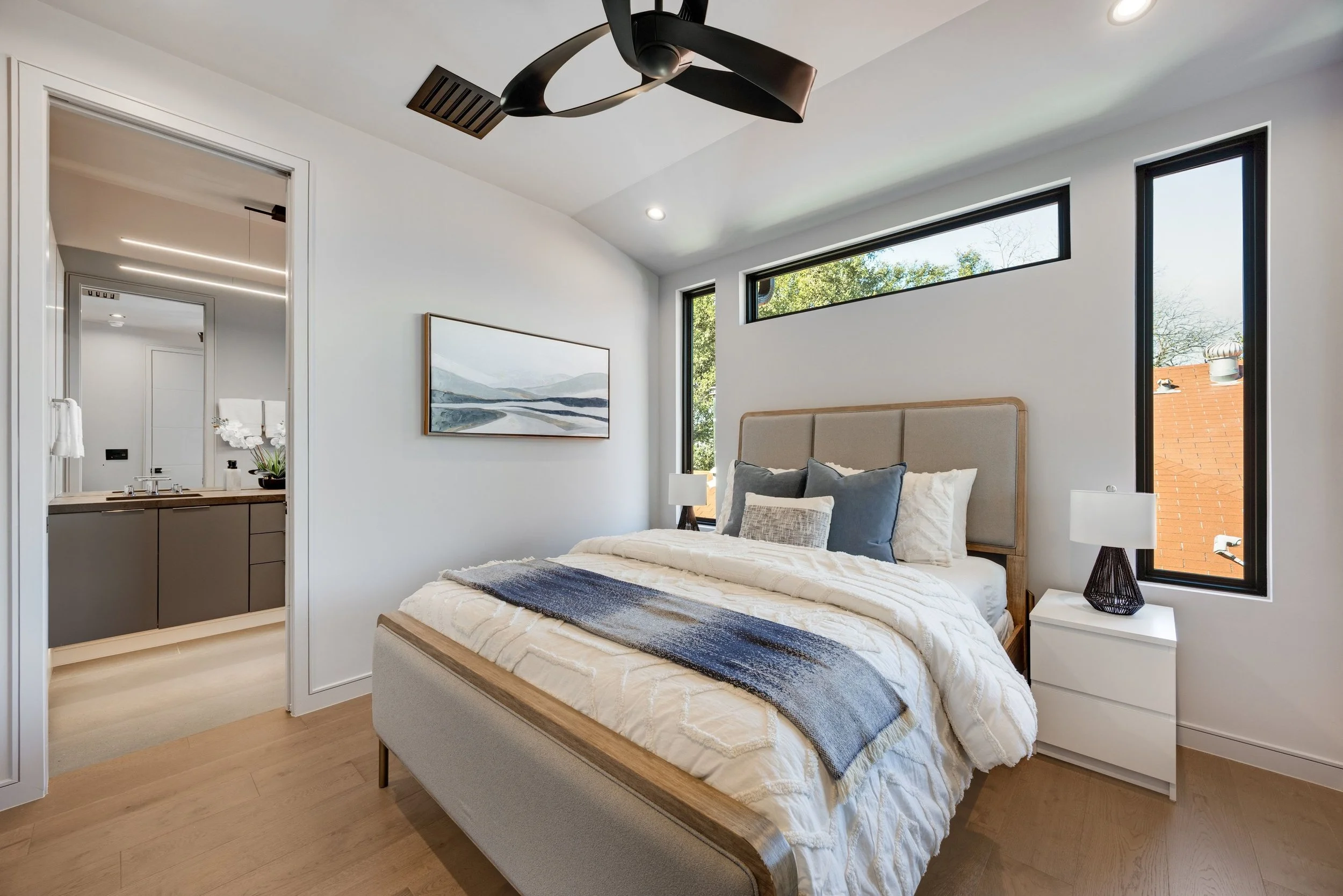 Modern bedroom with beige upholstered bed, white bedding, multiple pillows, two nightstands with lamps, large horizontal and vertical windows, and a ceiling fan. Part of an en suite bathroom with gray cabinets and a mirror is visible through an open 