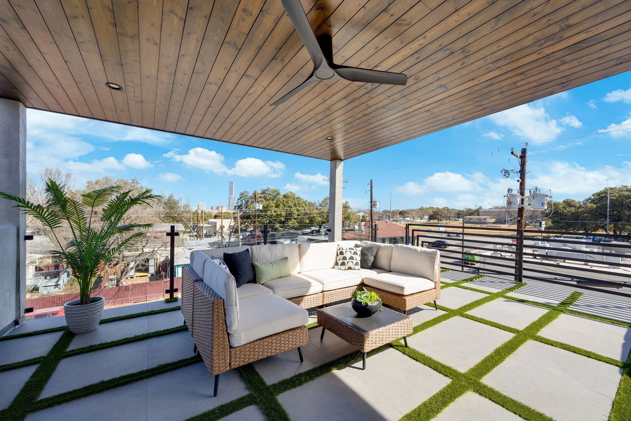 Balcony outdoor seating area with sectional sofa, coffee table, potted plant, and view of cityscape and power lines under a wooden ceiling with a ceiling fan.