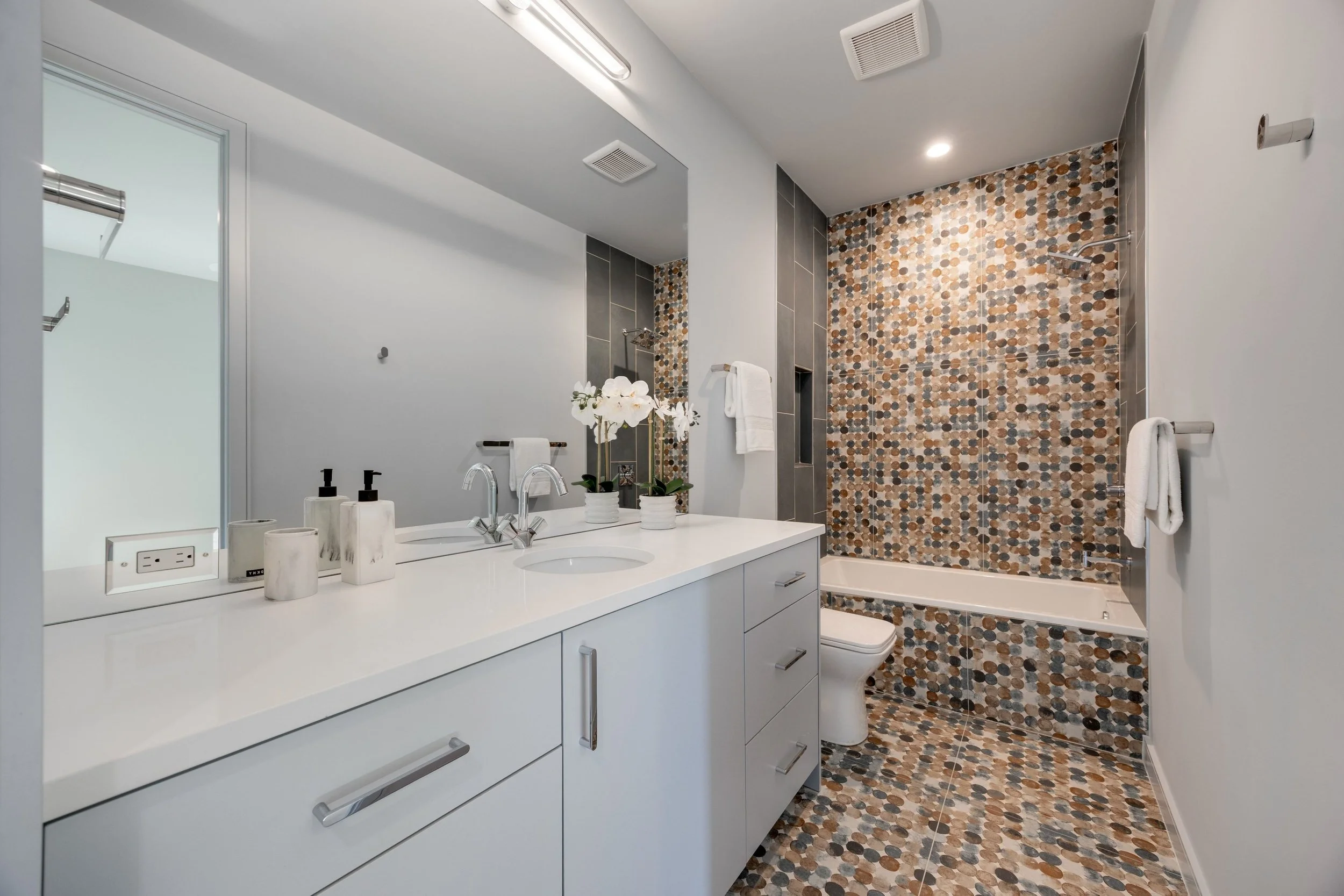 Modern bathroom with white vanity, marble accessories, plants, and a large mirror. The shower area features colorful pebble mosaic tiles on the wall and floor, with a built-in niche and hand towels hanging on chrome bars.