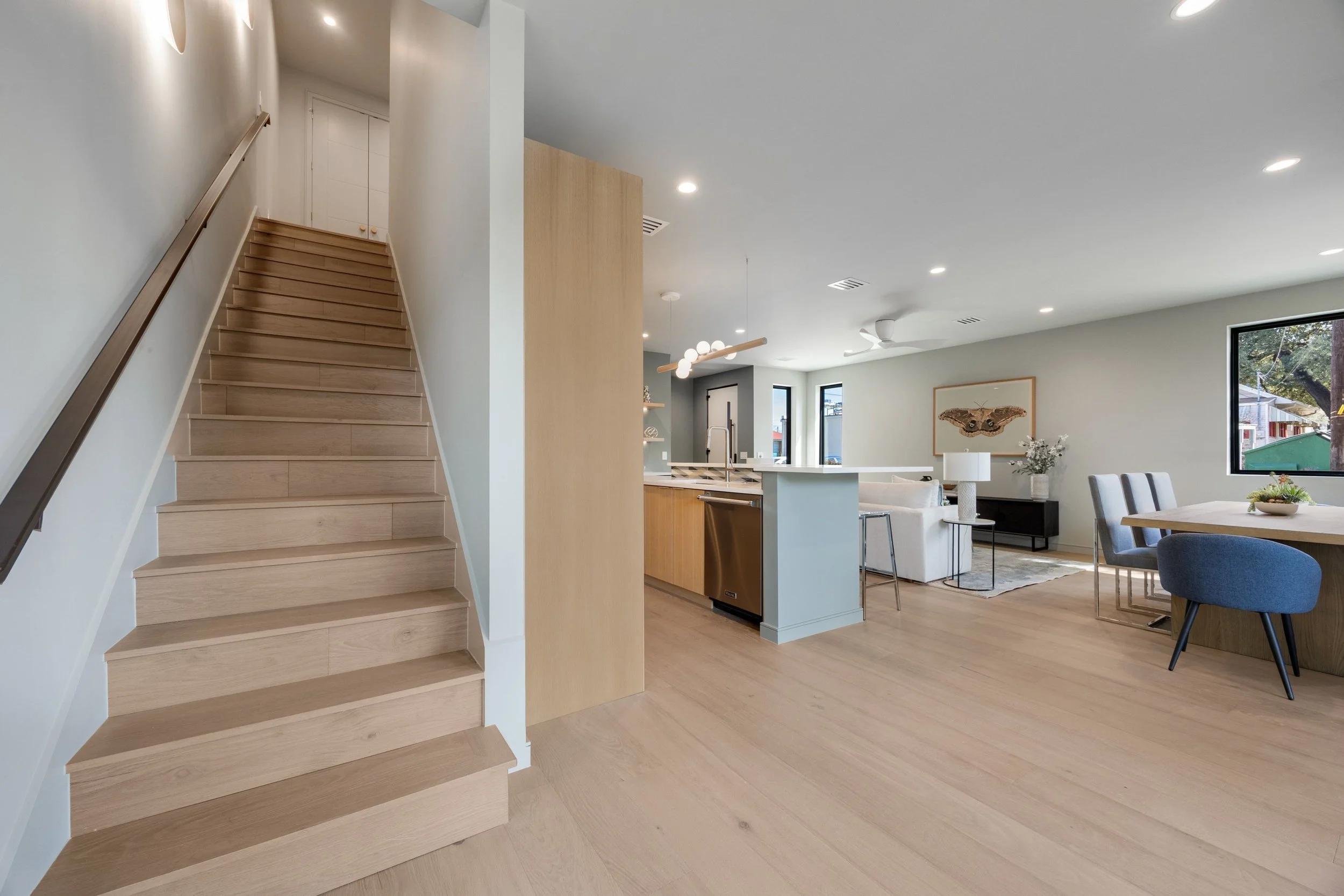 Open-concept modern apartment interior with light wood flooring, white walls, staircase, kitchen, dining, and living areas with large windows.