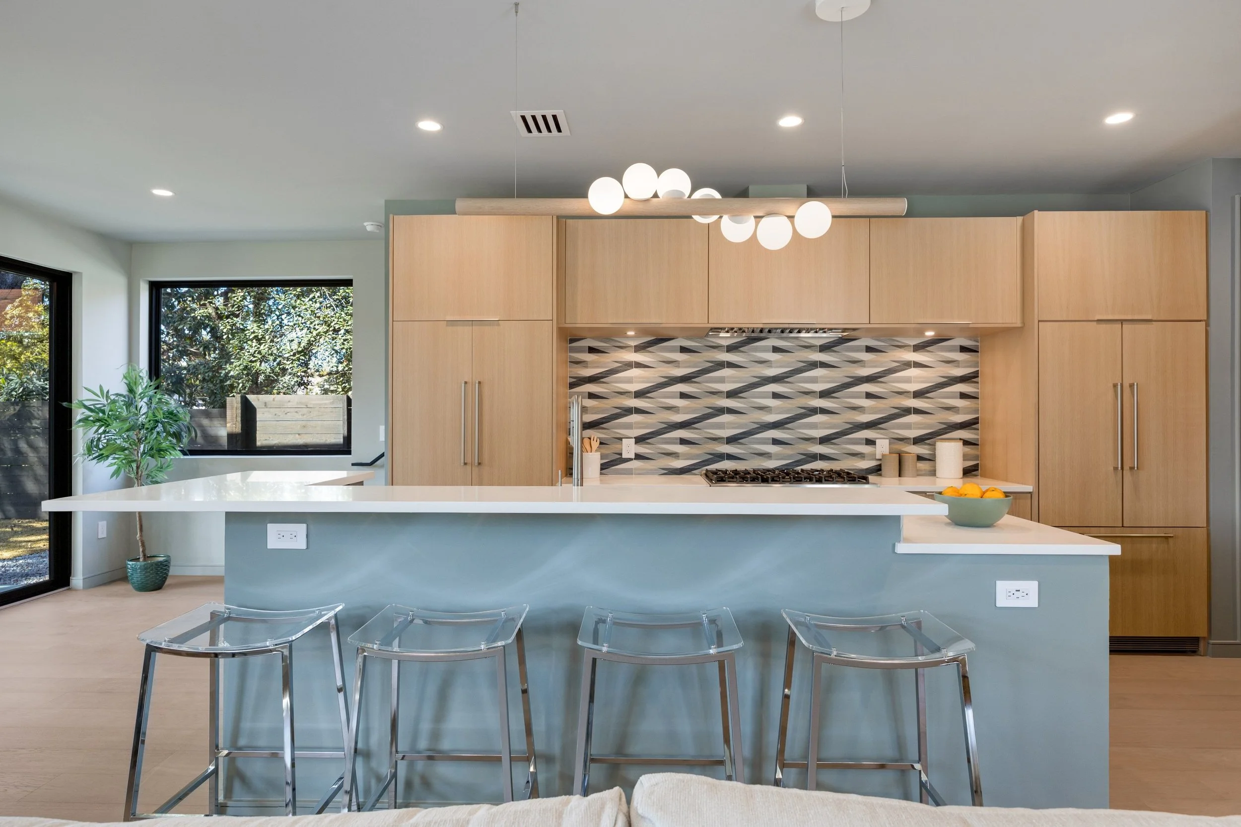 Modern kitchen with light wood cabinets, a geometric tiled backsplash, white countertops, and a center island with four clear plastic barstools. A bowl of oranges is on the island, and a potted plant is near large windows.