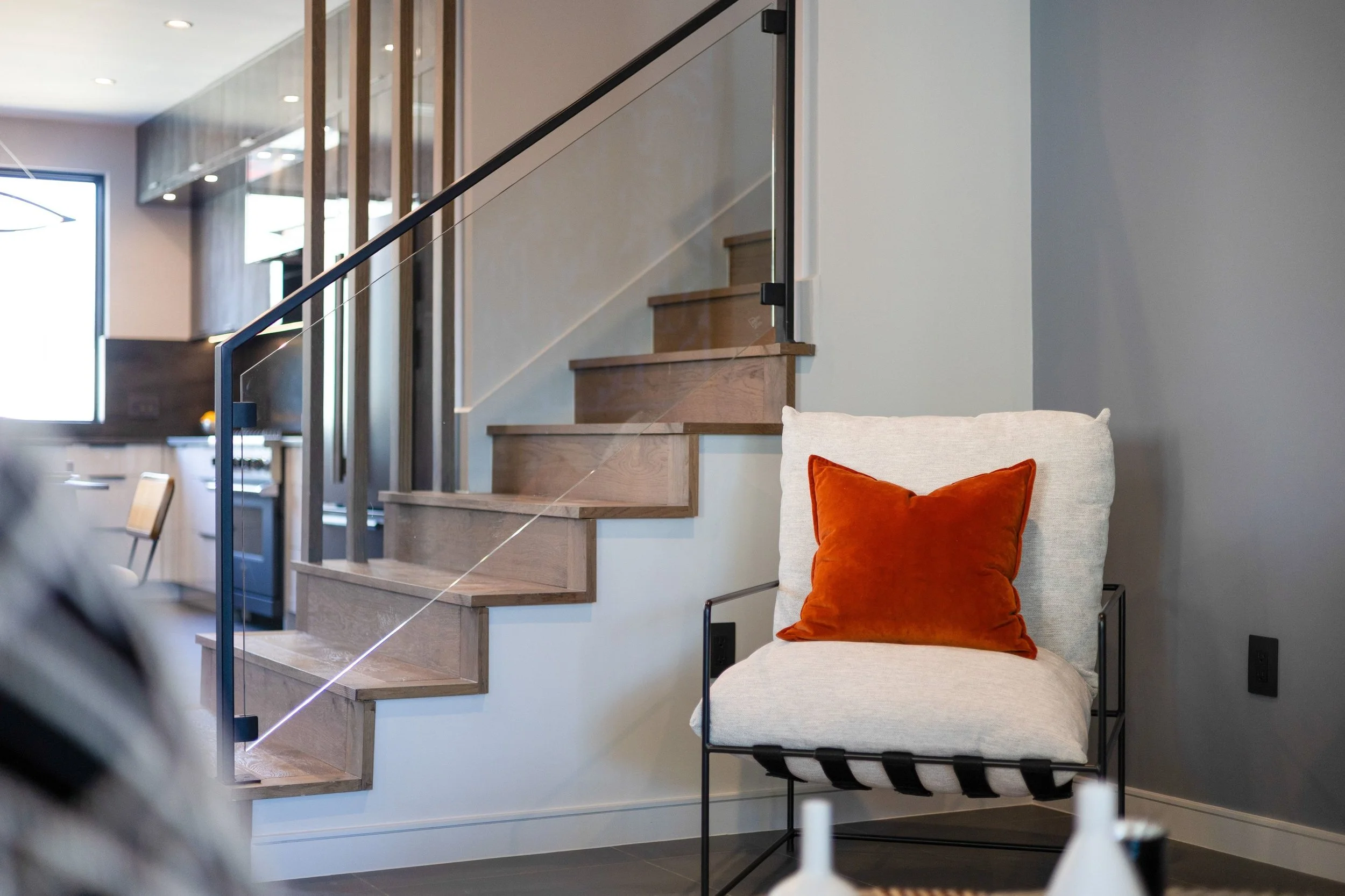 Living room corner with beige armchair and rust-colored throw pillow, wooden staircase with glass and black metal railing in the background, modern interior decor.