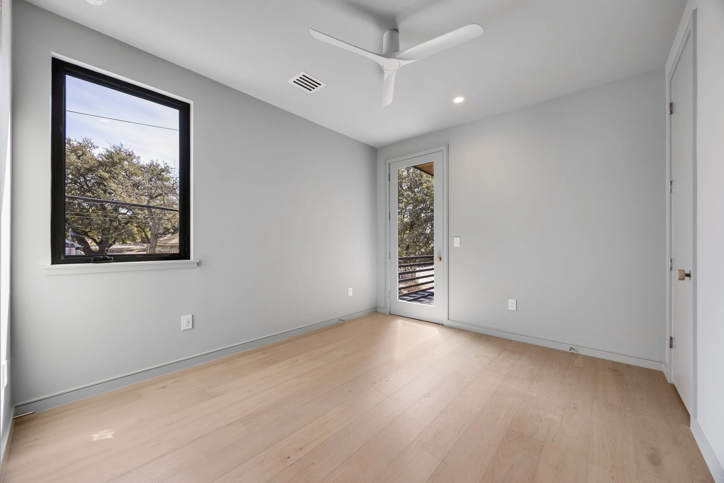 Empty room with a large window, a door leading to a balcony, light-colored wooden floors, white walls, and a ceiling fan.