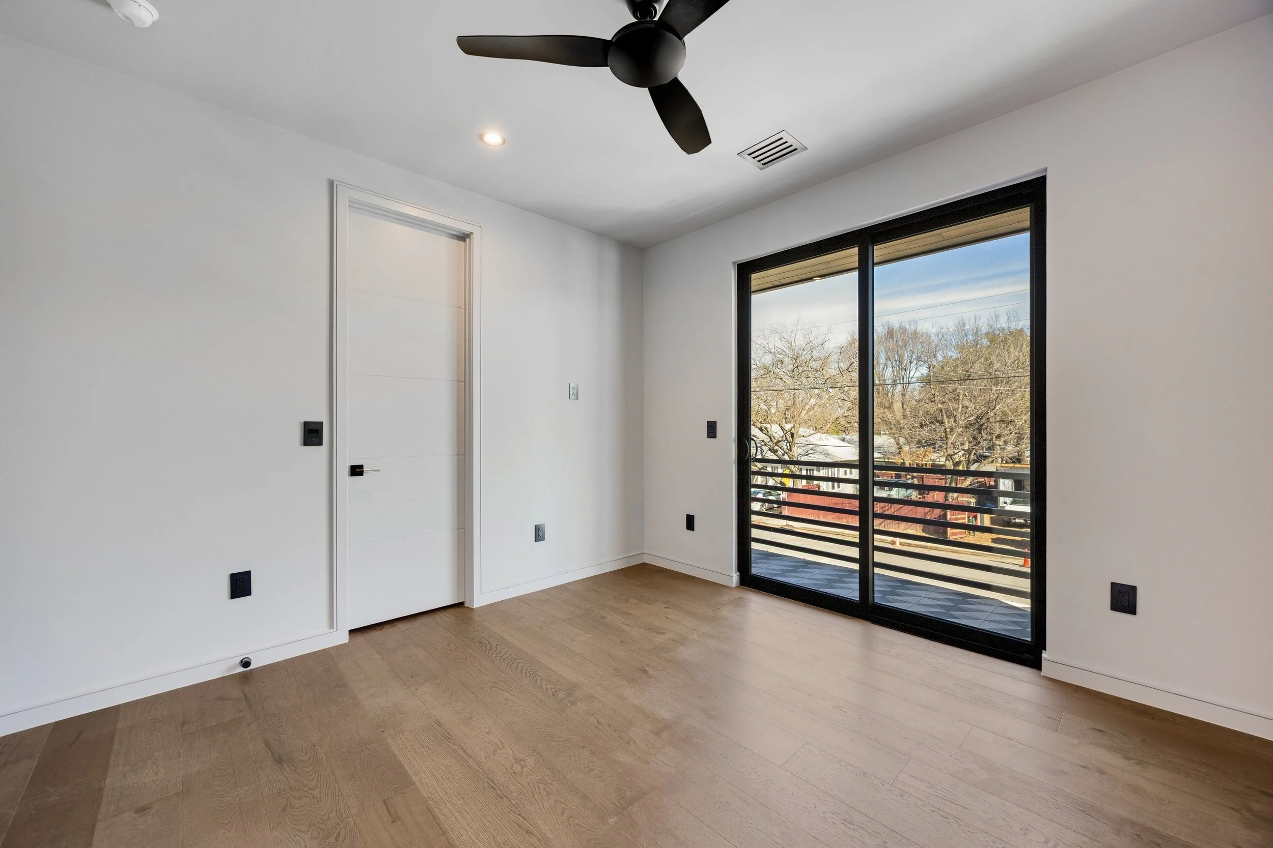Empty room with white walls, a wooden floor, and a sliding glass door leading to a balcony with a view of trees and rooftops outside.