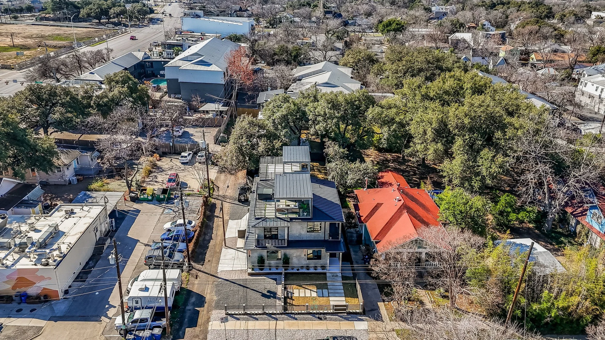 Aerial view of a neighborhood with a modern multi-story house, several smaller houses, trees, and parked cars.