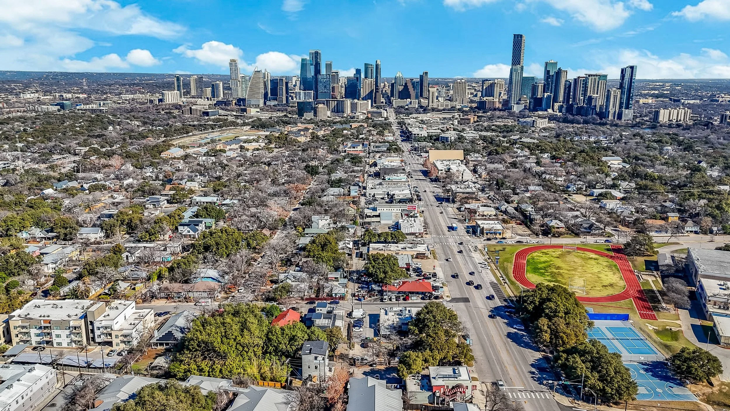 An aerial view of a cityscape with a downtown skyline featuring tall skyscrapers, a busy street, residential houses, a sports field with a running track, and tennis courts.