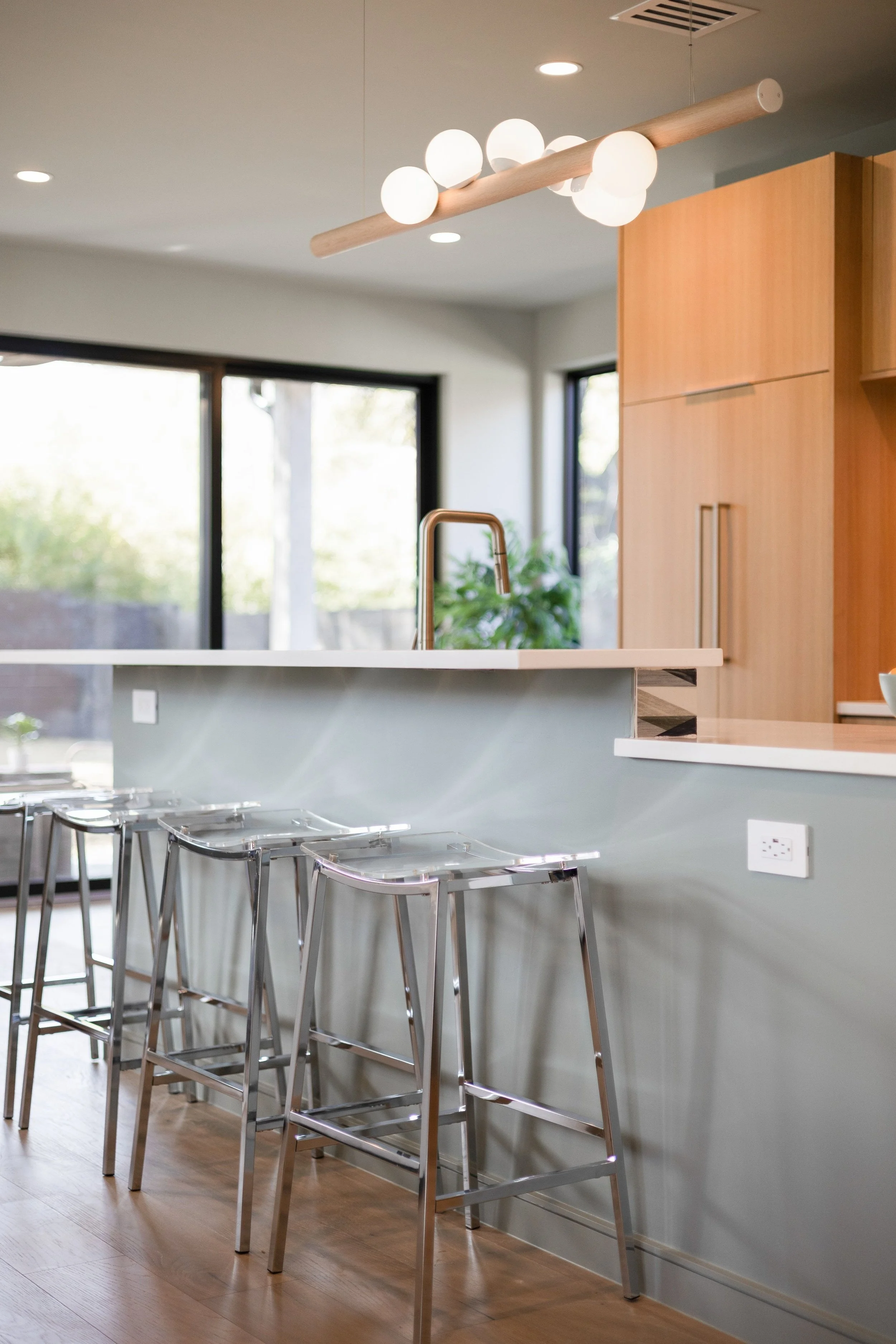 Modern kitchen with bar stools under a countertop, pendant light fixture with white globes, large windows, and wooden cabinets.