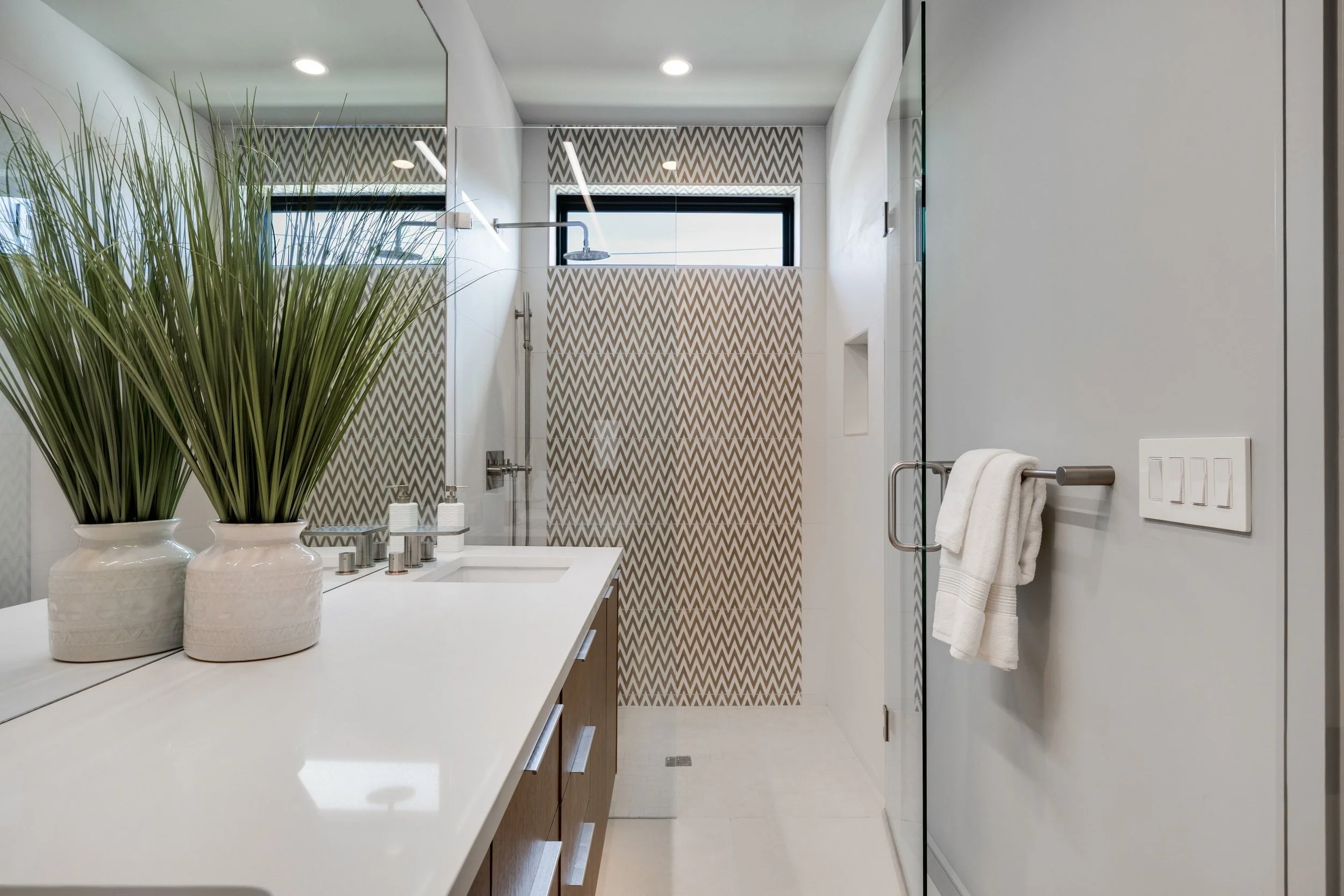 A modern bathroom with a double vanity countertop holding two potted green plants, a sink, and a mirror. In the background, there is a walk-in shower with a patterned shower wall, a window with a geometric valance, and a towel hanging on a rod.