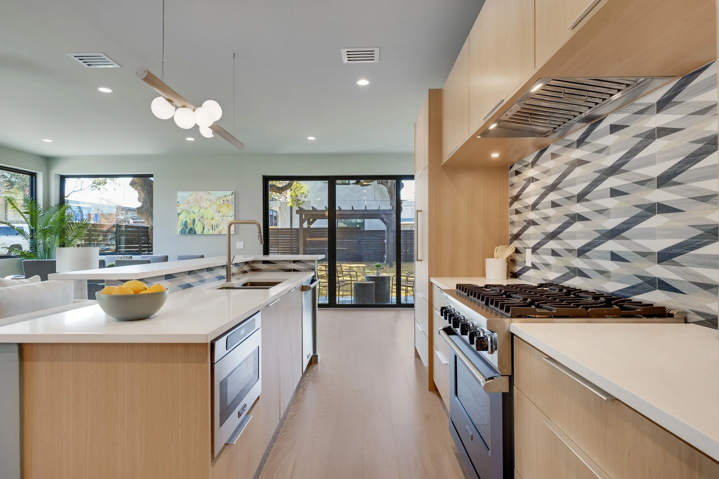 Modern kitchen with light wood cabinetry, geometric backsplash, stainless steel appliances, and a large white island with a bowl of lemons, overlooking a living area and backyard through sliding glass doors.