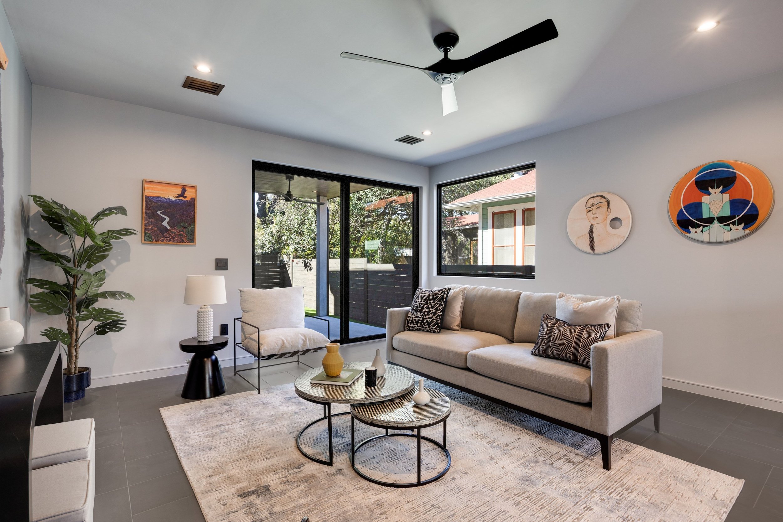 Modern living room with beige sofa, coffee tables, artwork, large windows, sliding door leading outside, ceiling fan, and potted plant.