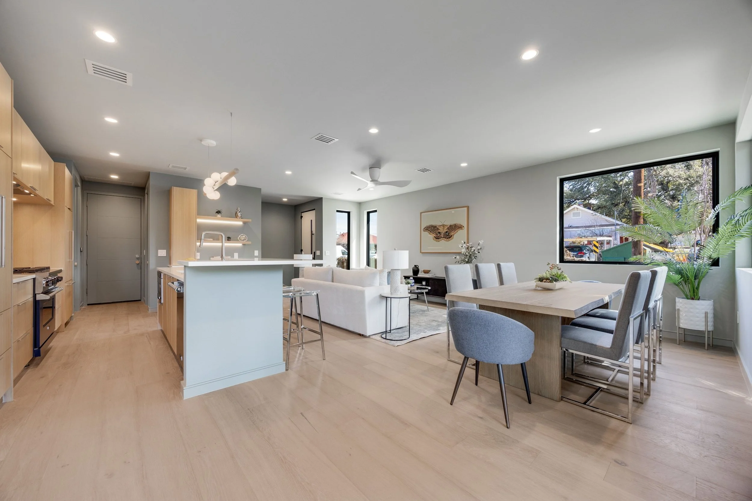 Open-concept living room and kitchen with light wood flooring, white walls, modern furniture, and large windows showing outdoor scenery.