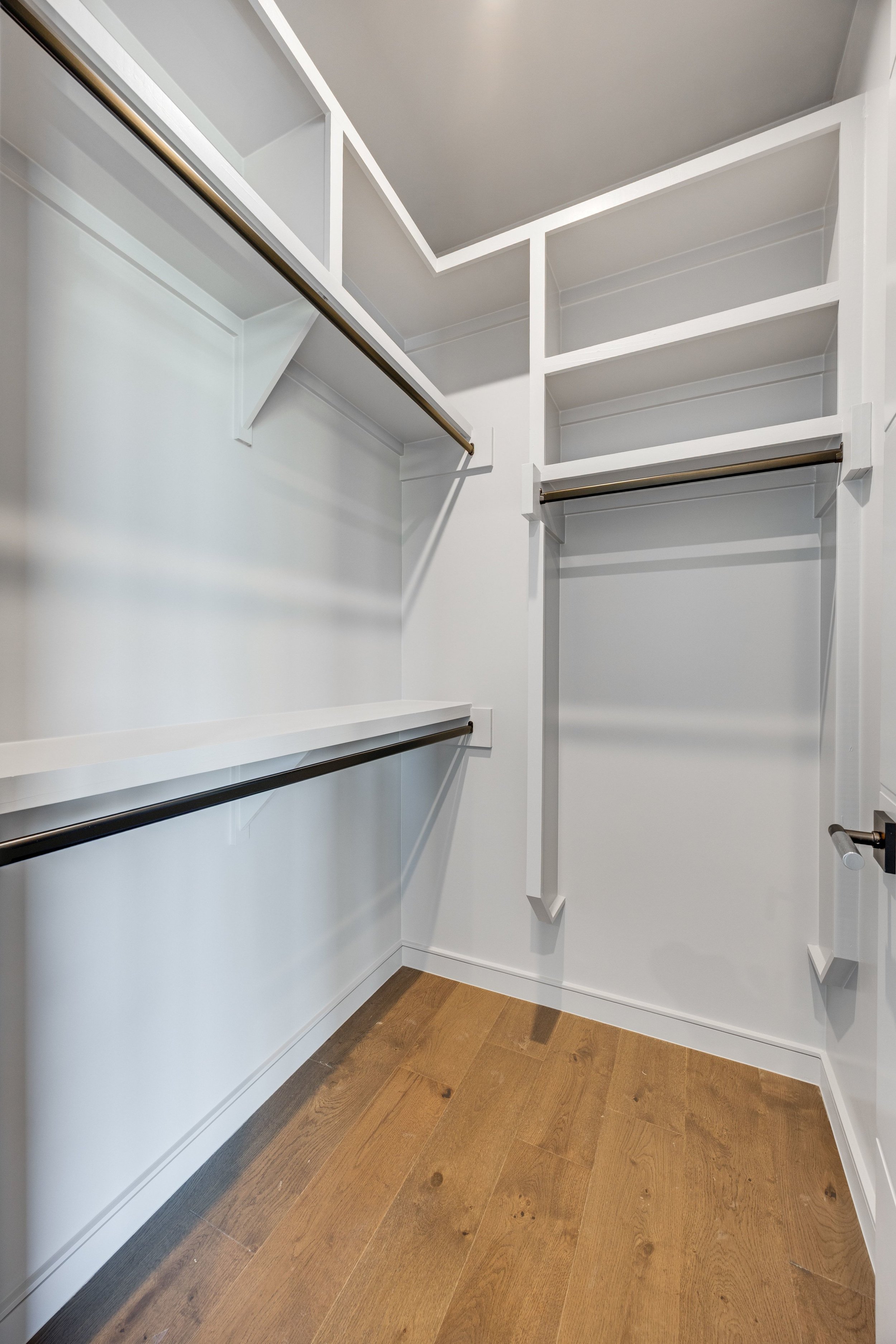 Empty walk-in closet with white shelves and hanging rods, a wood floor, and a door handle visible on the right.