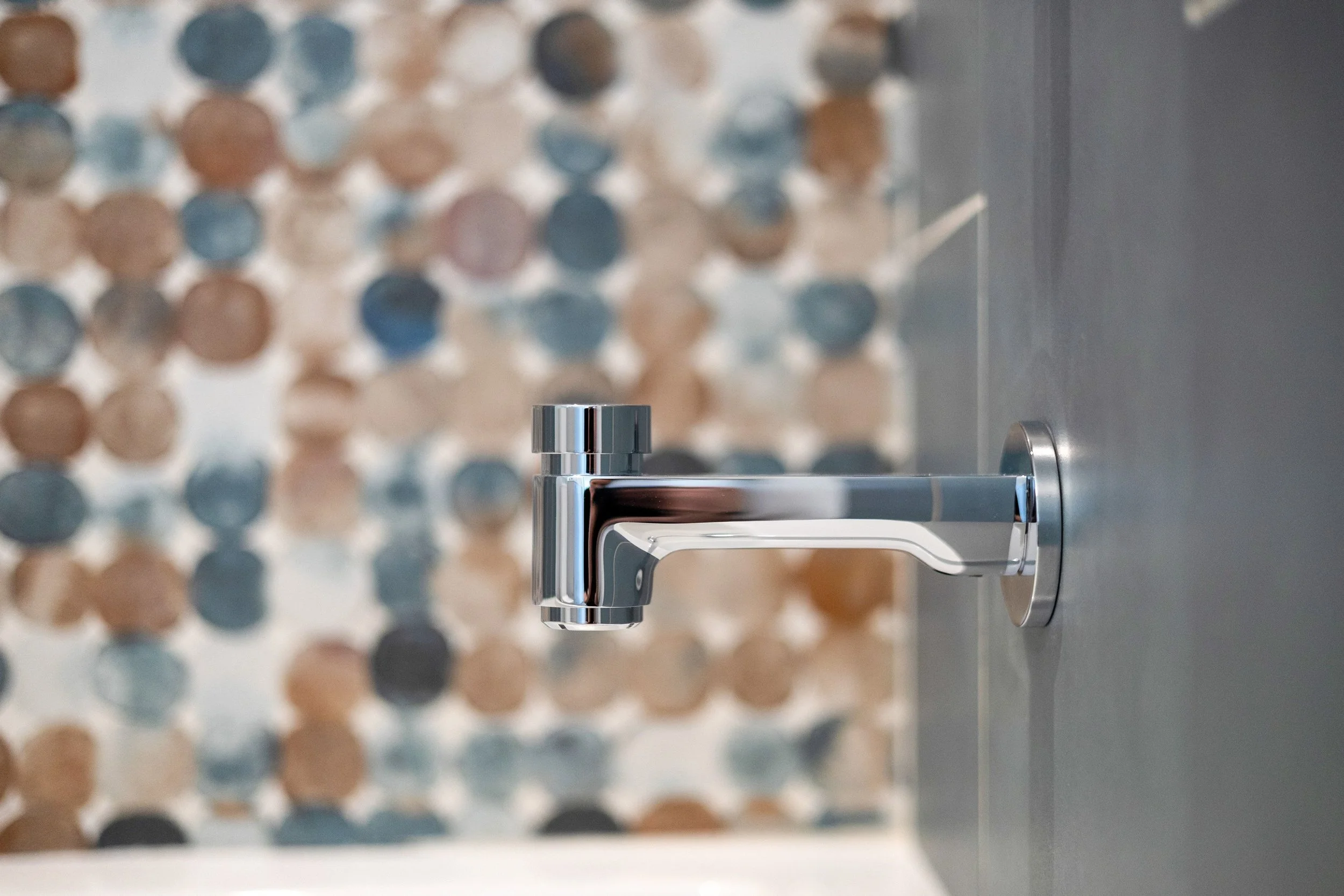 A chrome-finished faucet mounted on a bathroom wall with a mosaic tile background composed of round tiles in shades of brown, gray, and blue.