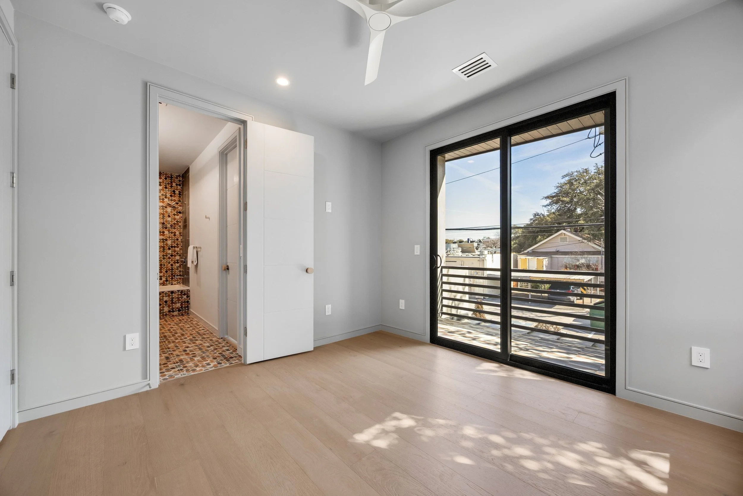 Empty room with light wood flooring, white walls, sliding glass door leading to balcony, and an open door revealing a bathroom with colorful mosaic tiles.