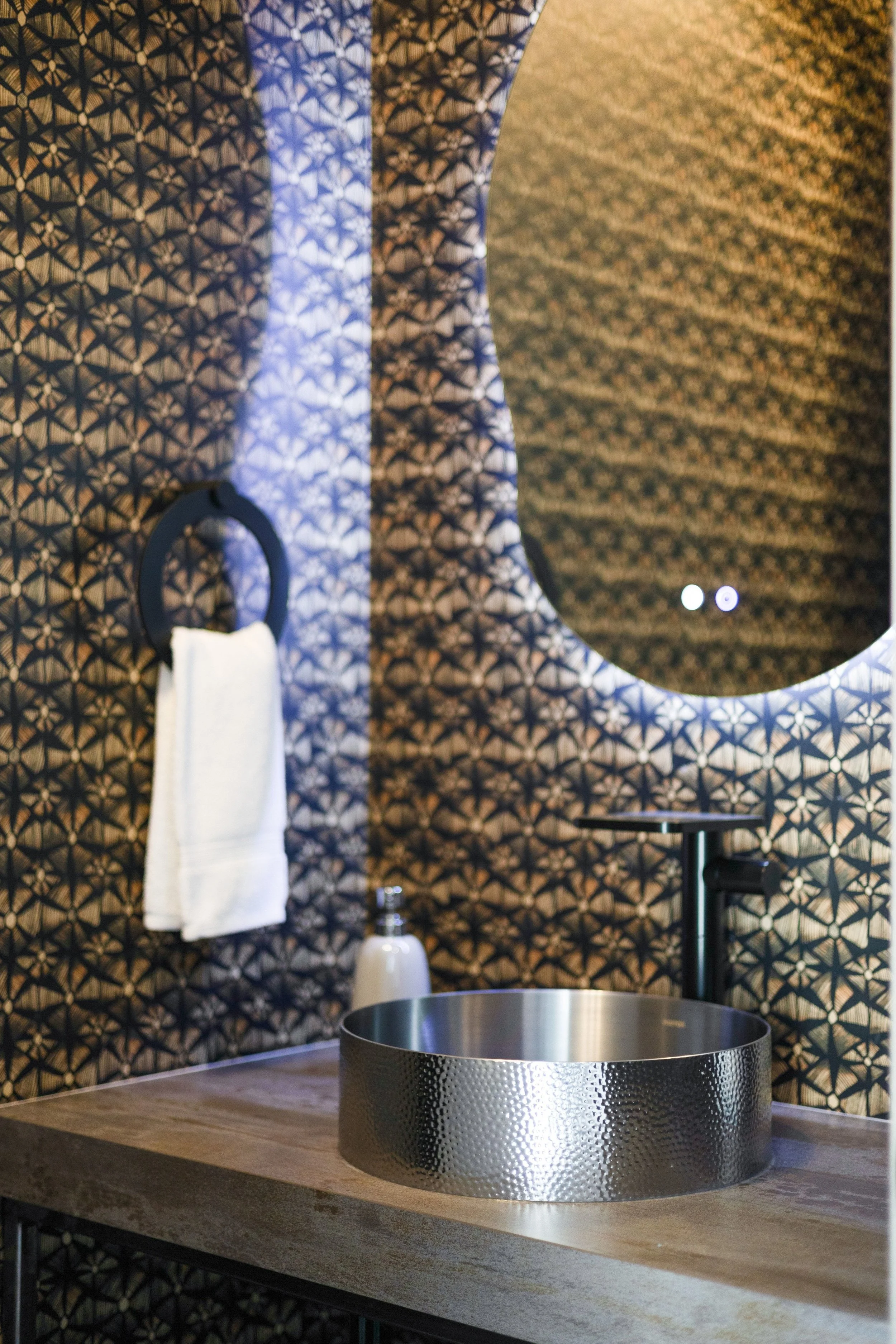 Modern bathroom with a textured metal basin sink, a mirror with a geometric patterned frame, a towel ring with a white towel, a soap dispenser, and wallpaper with a black and gold geometric design.
