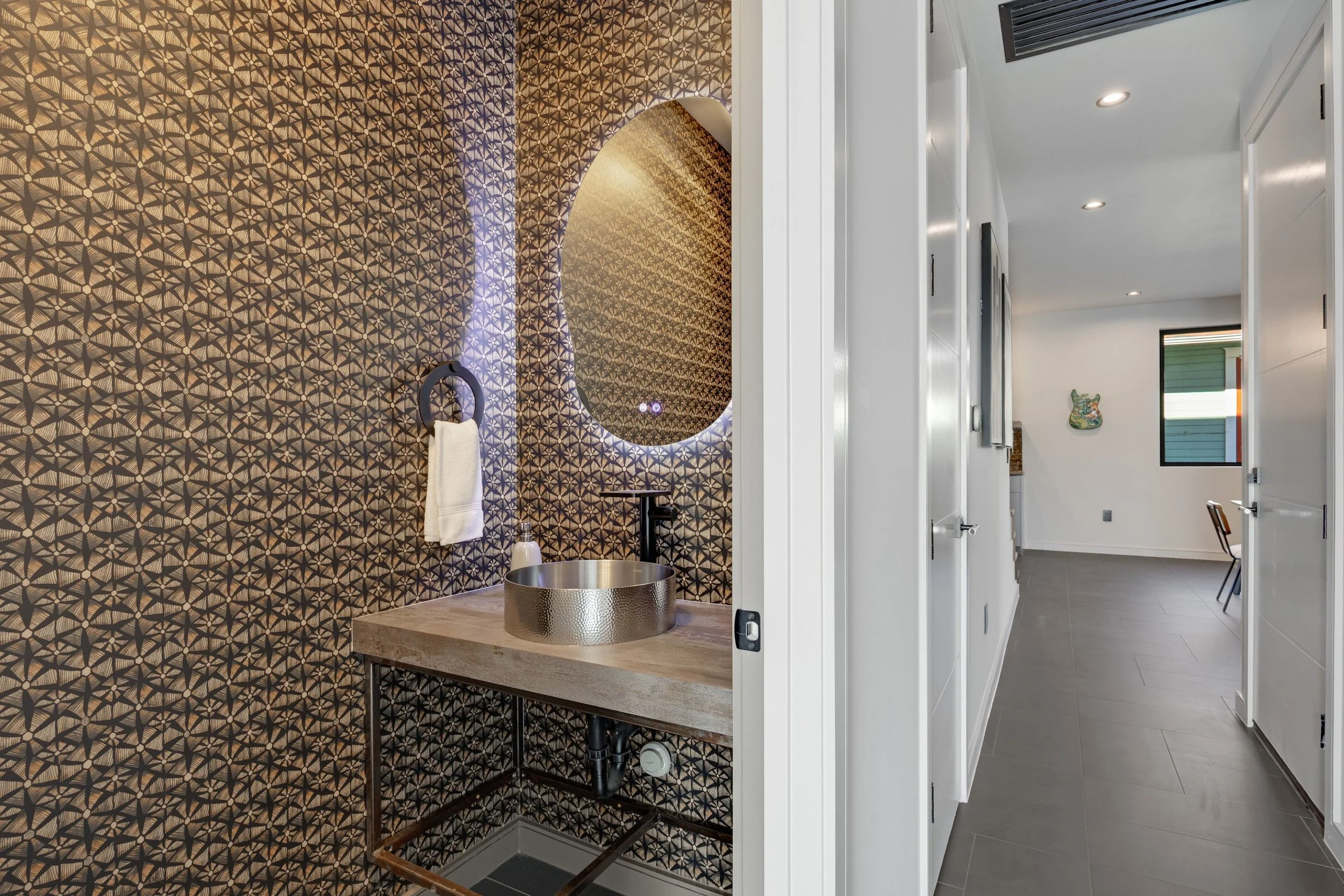 An interior view of a modern bathroom and hallway, featuring geometric patterned wallpaper in a bathroom with a gold circular sink, a mirror with lighting, and a hallway with white walls, gray flooring, and a window with green and white blinds.