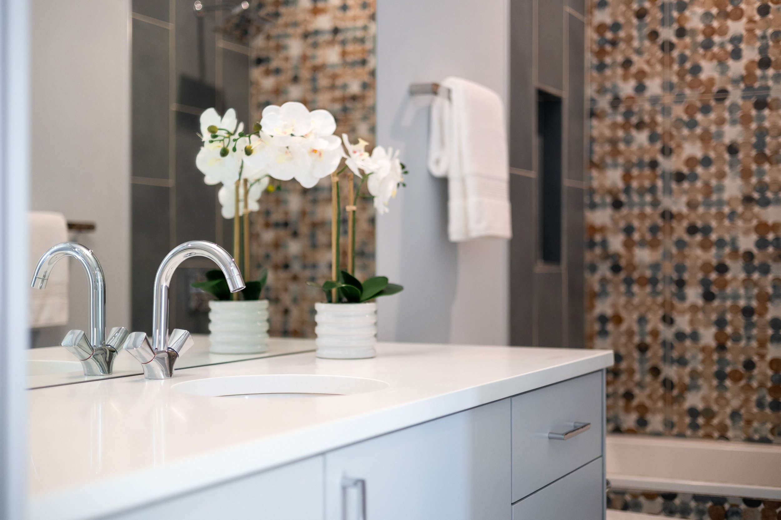 A modern bathroom featuring a white countertop with a sink, chrome faucets, and white orchid plants in striped pots, with a background of a patterned tile wall and a towel hanging on the wall.