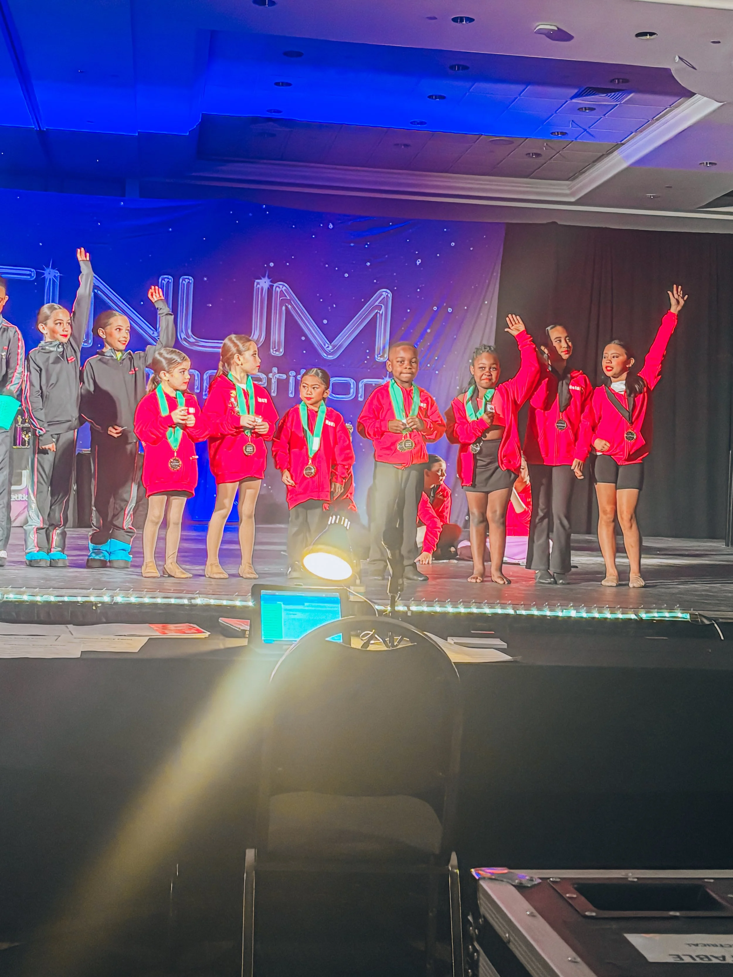 Children in red and black uniforms standing on a stage during an awards ceremony, with medals around their necks and a blue-lit backdrop that reads 'NAML'.