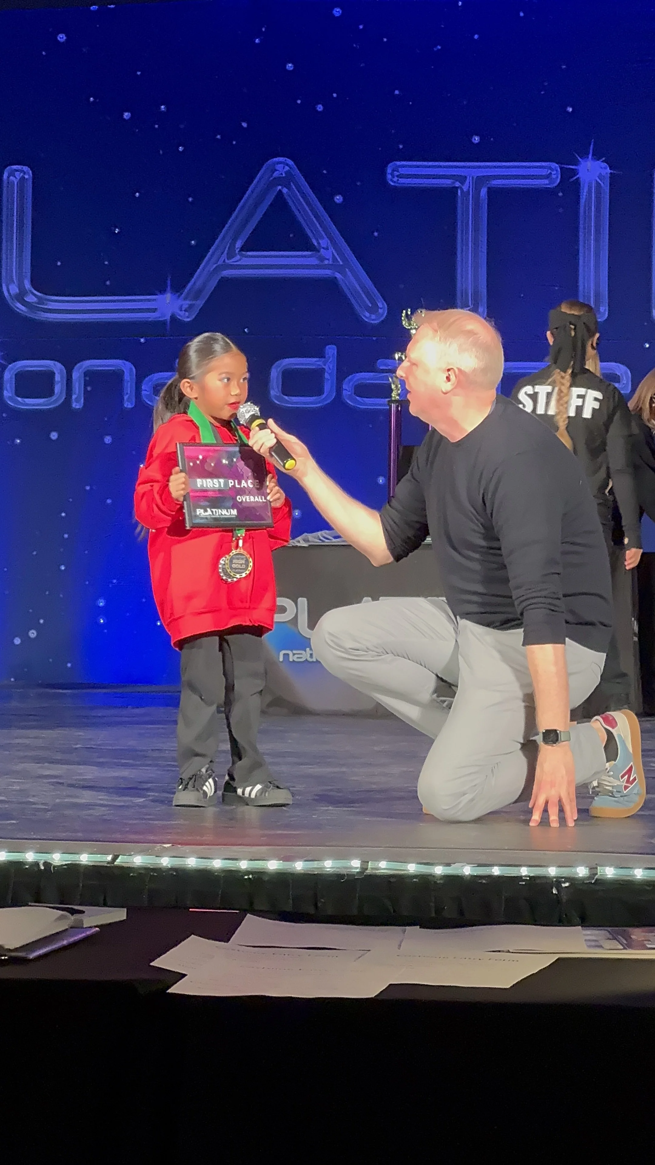 A young girl in a red jacket holding a first-place award medal and a plaque, being interviewed on stage by a man kneeling on one knee, at a dance competition event. She is wearing black shoes with white stripes, and there are staff members and a large blue backdrop with the word 'LATIT' visible behind them.