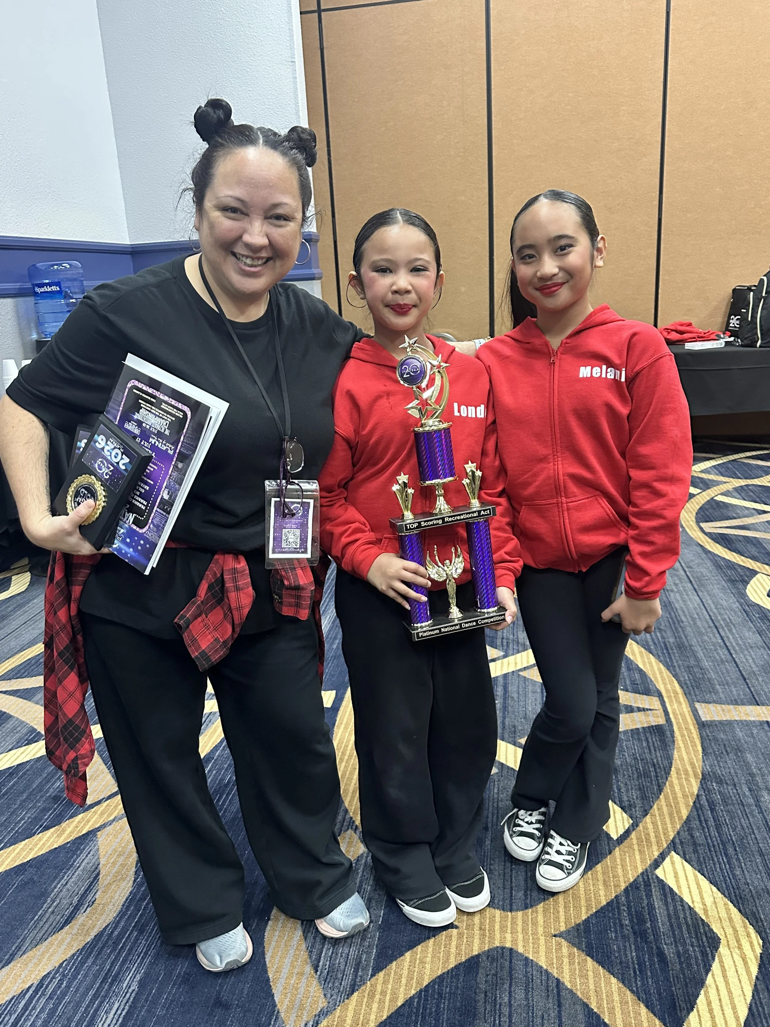 Three females, two young girls and an adult woman, standing together indoors, smiling at the camera. The girl in the middle is holding a large trophy. The woman and the girl on the right are wearing red hoodies, and the girl on the left is wearing a red top. They appear to be at a competition or award event.