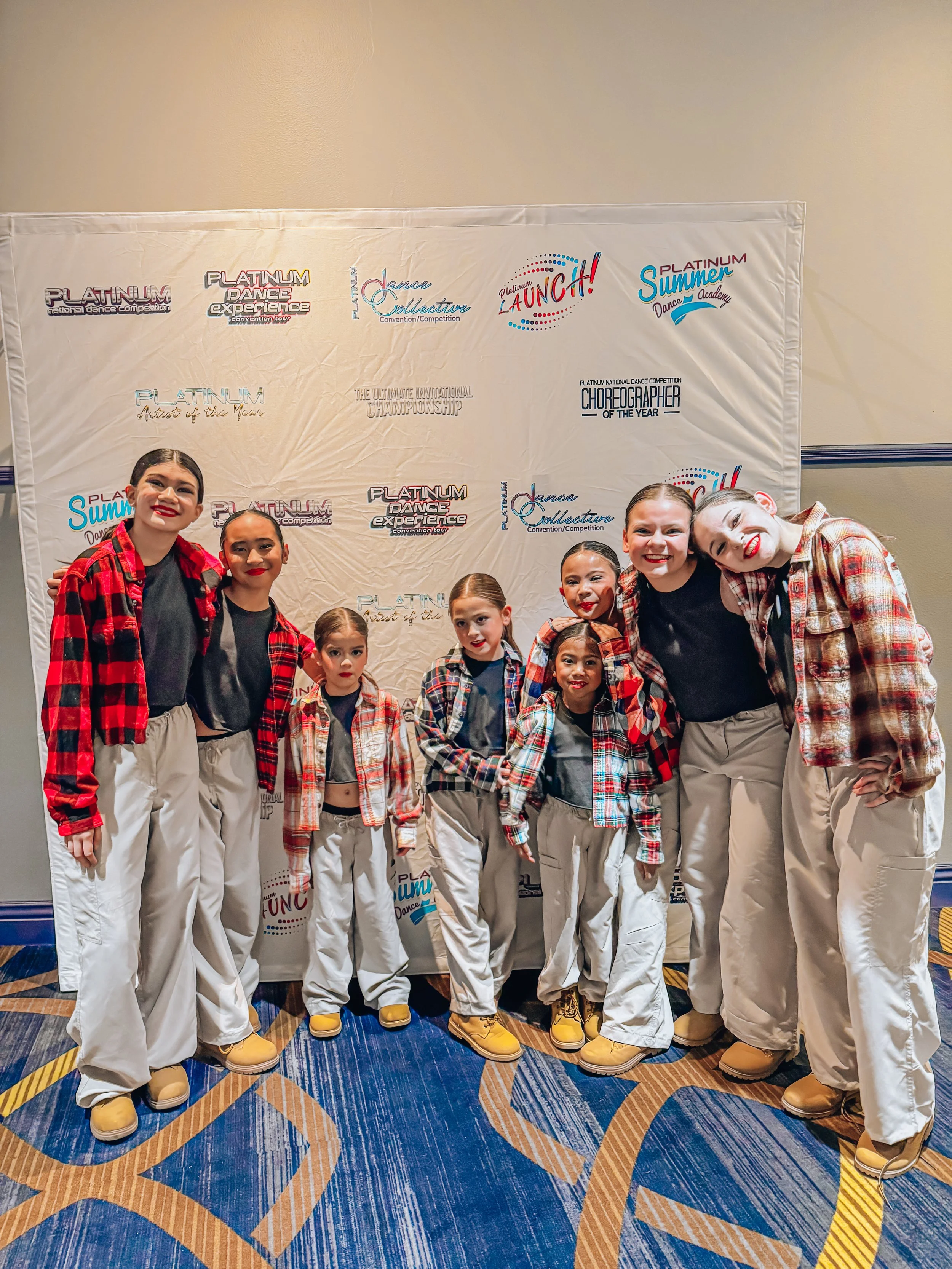 Group of young female dancers in plaid shirts, black tops, beige pants, and dance shoes standing together at a dance competition backstage