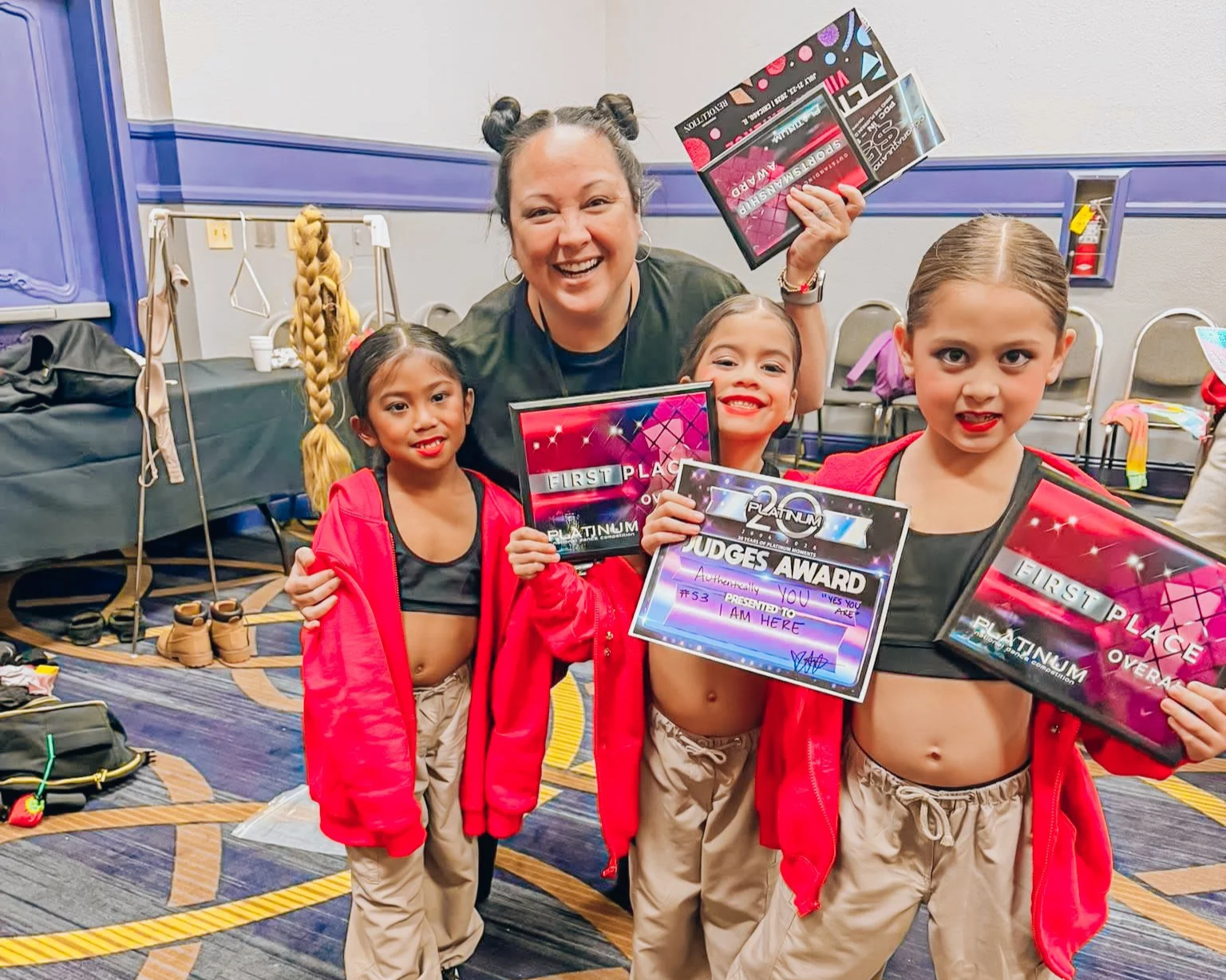 Four young girls and a woman celebrating an award at a dance competition, holding plaques and a judges award, in a room with chairs, shoes, and bags in the background.