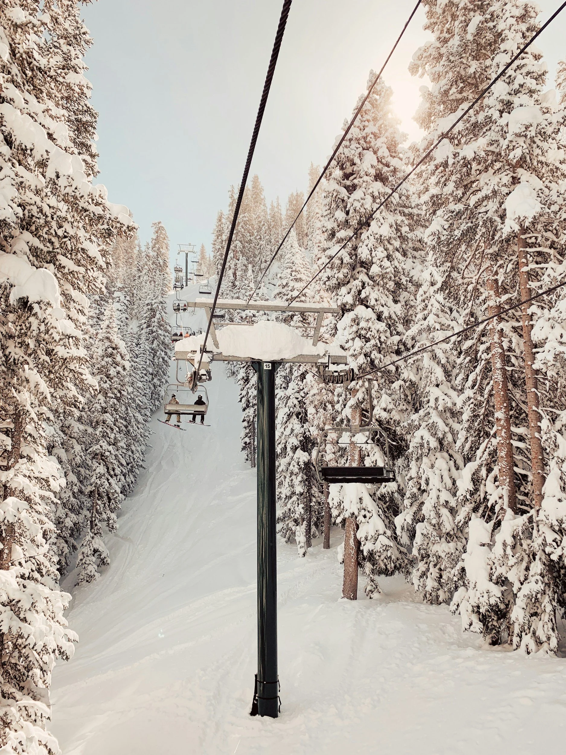 Ski lift carrying people up a snowy mountain surrounded by snow-covered pine trees with sunlight filtering through.