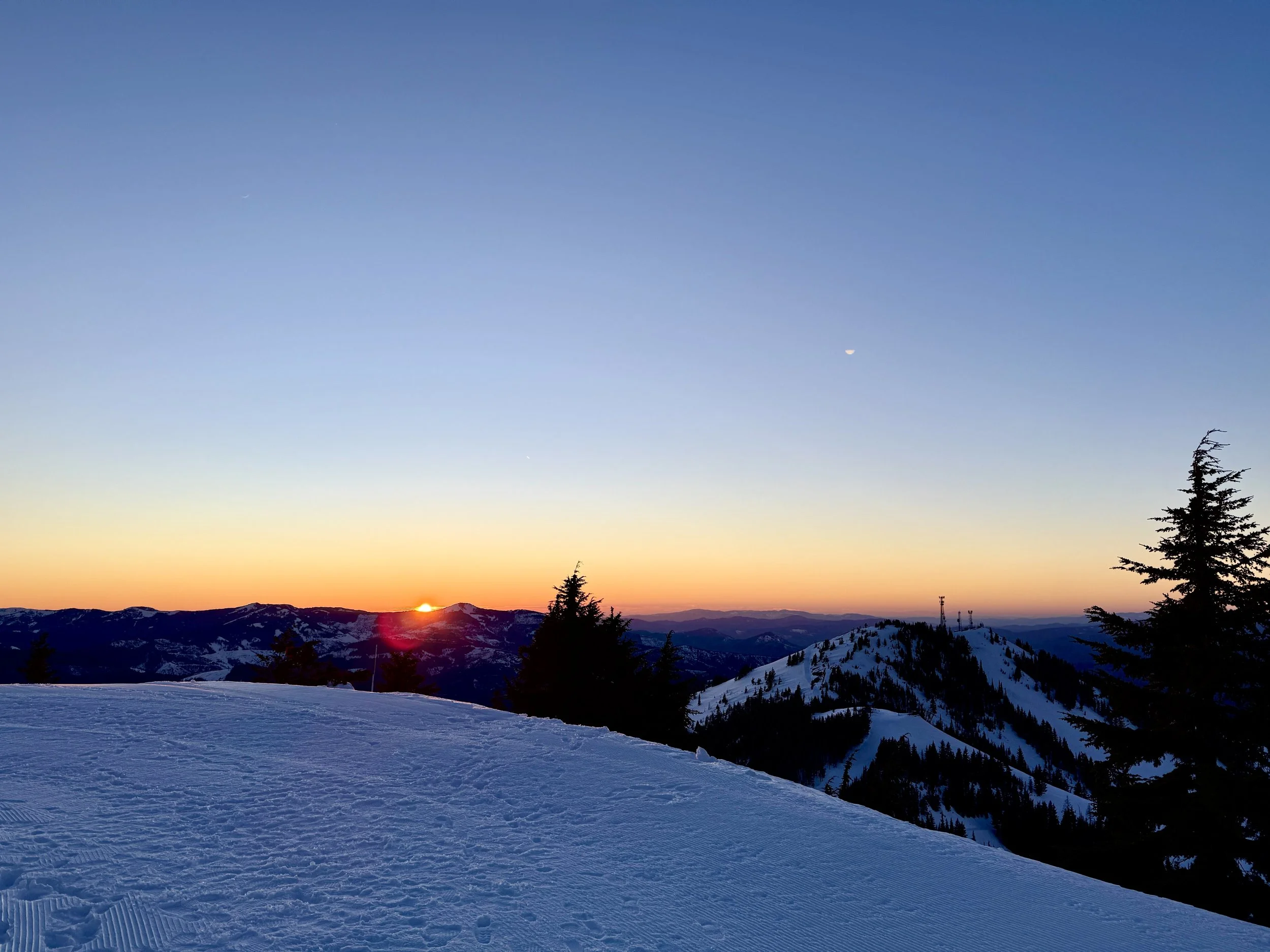 Sunset over snow-covered mountain landscape with evergreen trees and clear blue sky in the background.