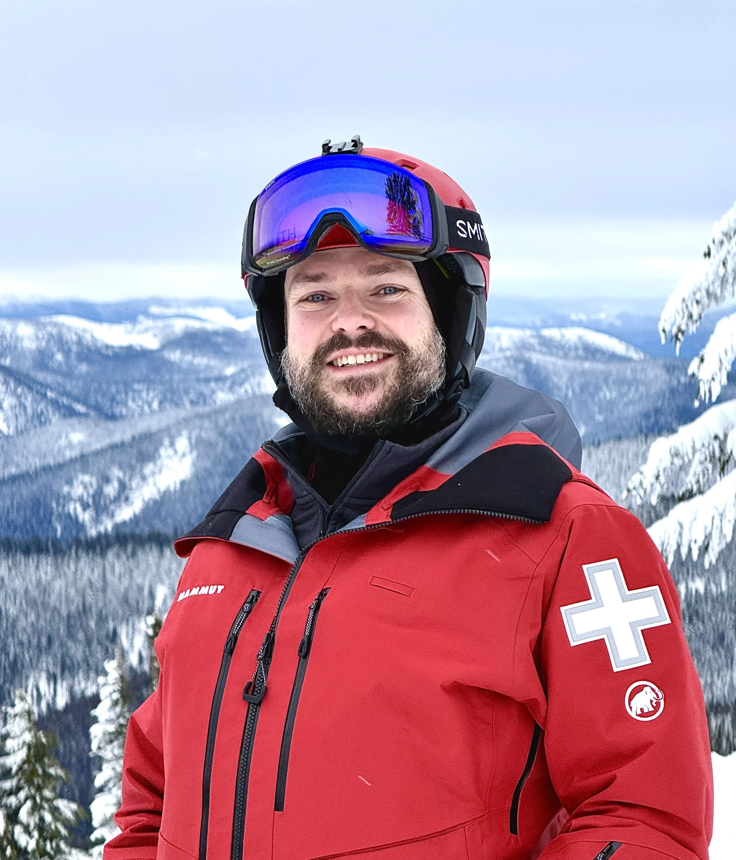 Man in red ski jacket, helmet, and goggles standing in snowy mountainous landscape.