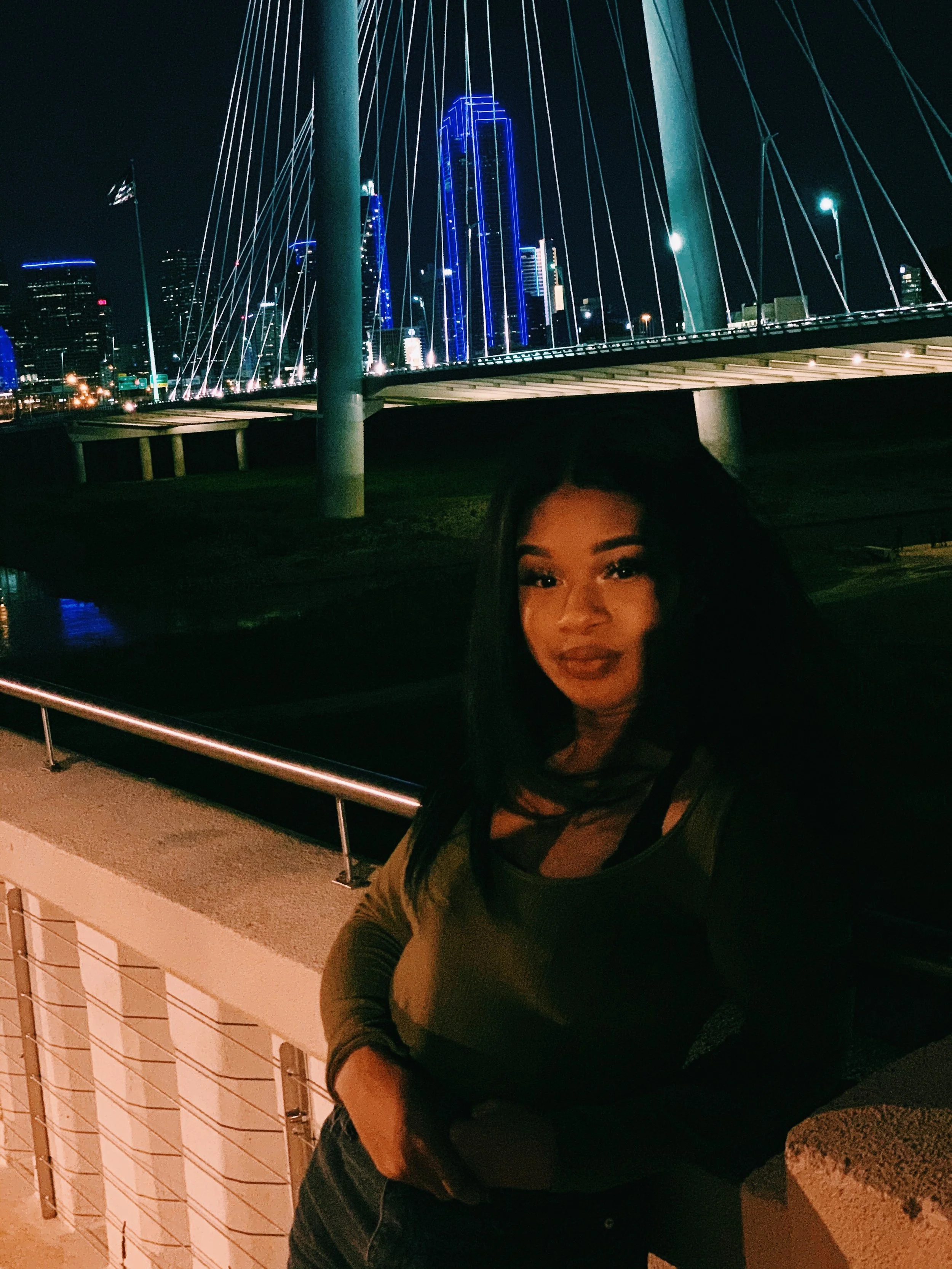 A woman standing outside at night, with a city skyline and illuminated bridge behind her.