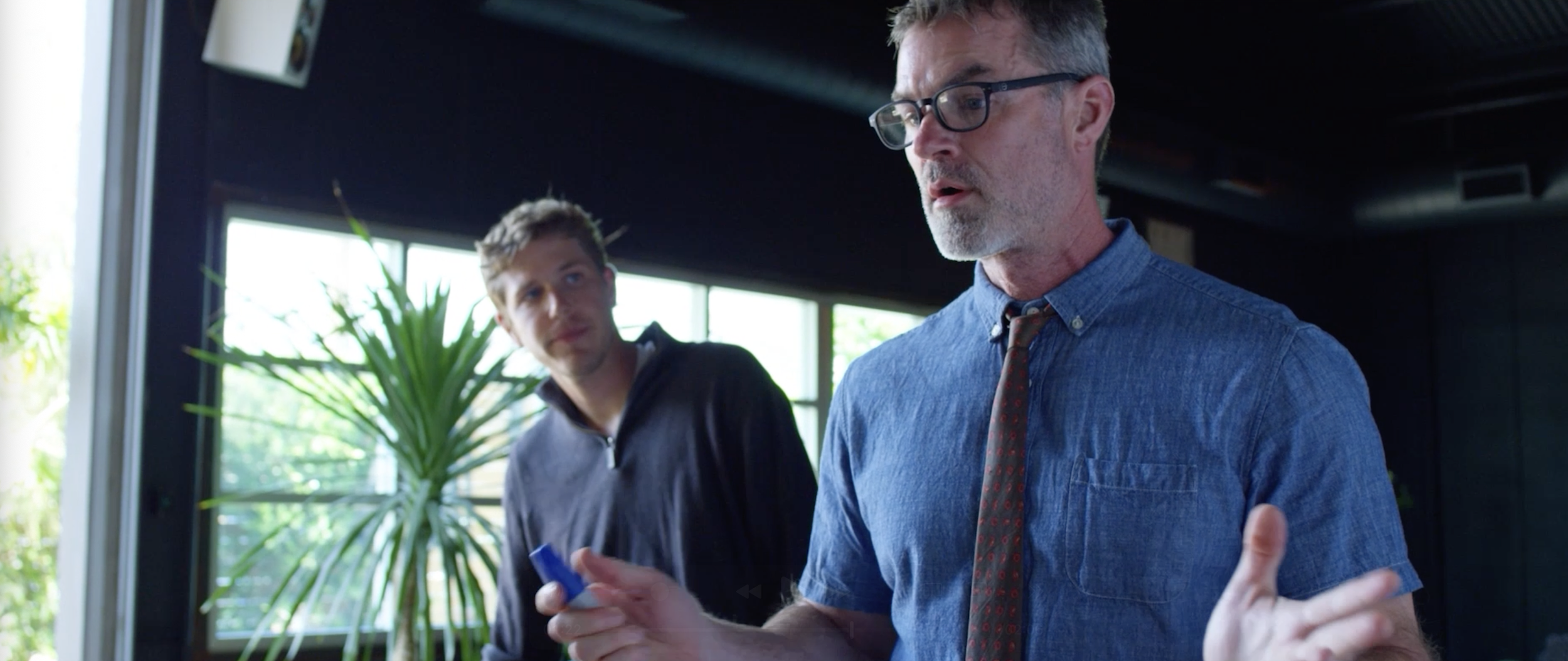 Two men in a discussion inside a room with large windows and a potted plant. One man wears glasses and a blue shirt, gesturing with his hand; the other man stands nearby, holding a small object, looking at him.