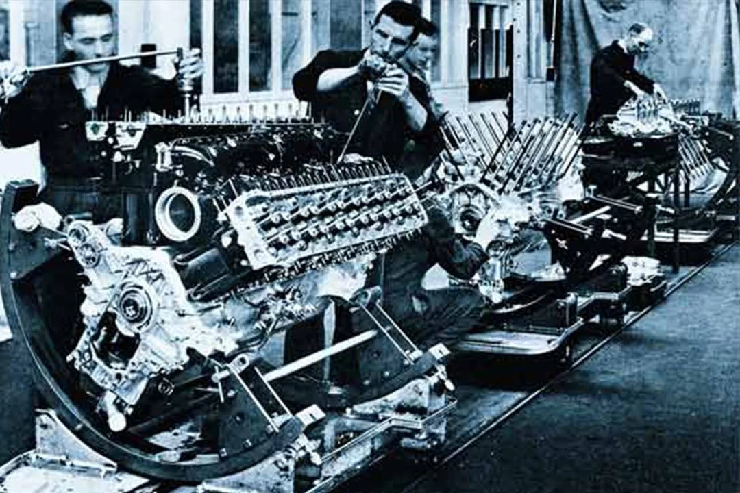 Three men working on an aircraft engine, with one of them playing a harmonica, in black and white.