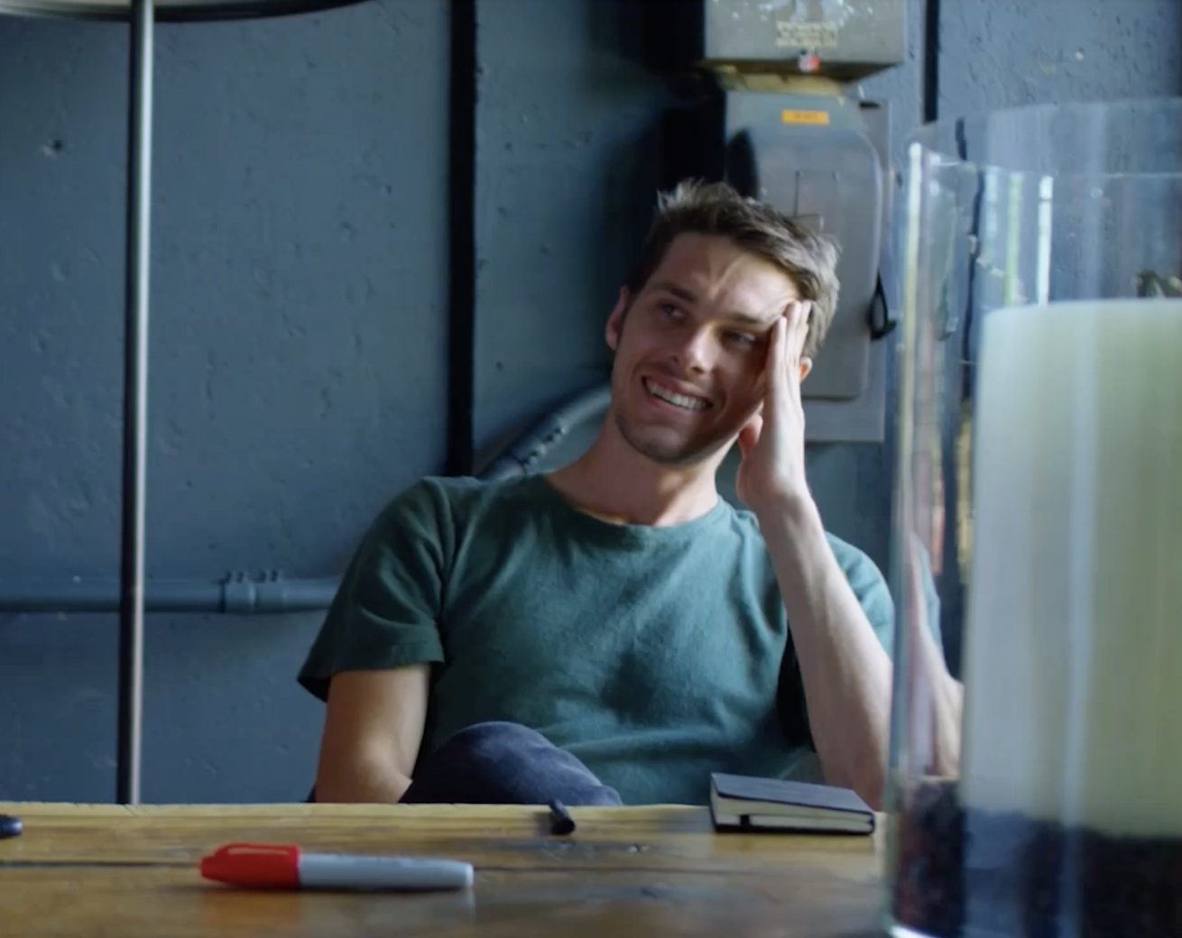 A young man with short, light brown hair smiling and resting his head on his hand while sitting at a desk in front of a computer. There are pens and a notebook on the desk, and the background features a plain, dark gray wall with equipment mounted to it.