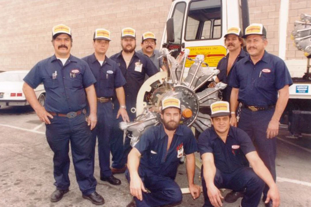 Group of seven men, wearing blue uniforms and yellow hats, standing in front of a large engine or machinery, with a truck and a brick wall in the background.