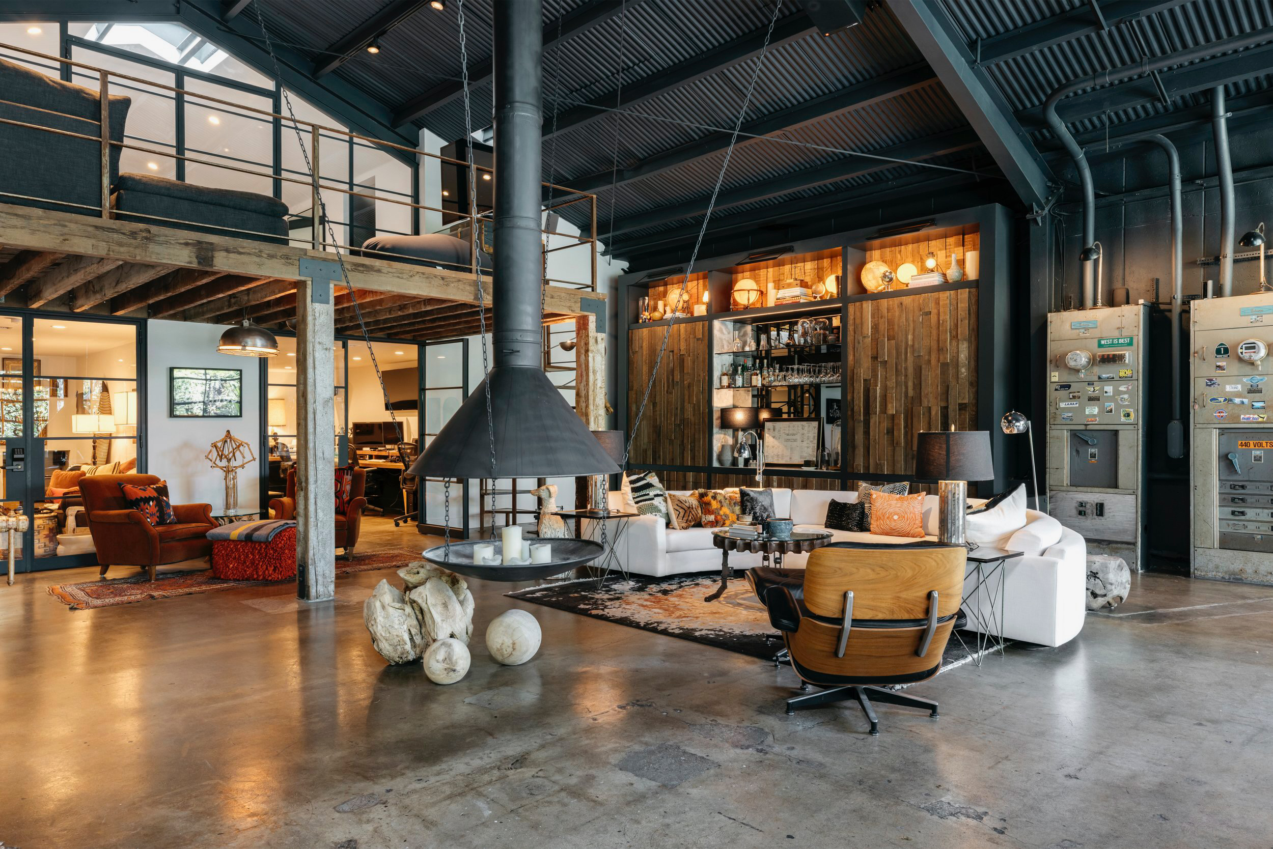 Modern industrial-style living room with lofted second floor, featuring exposed metal beams, wooden wall panels, a bar area, and various seating options including a white sofa, wooden chairs, and armchairs, with decorative elements like pillows, lamp