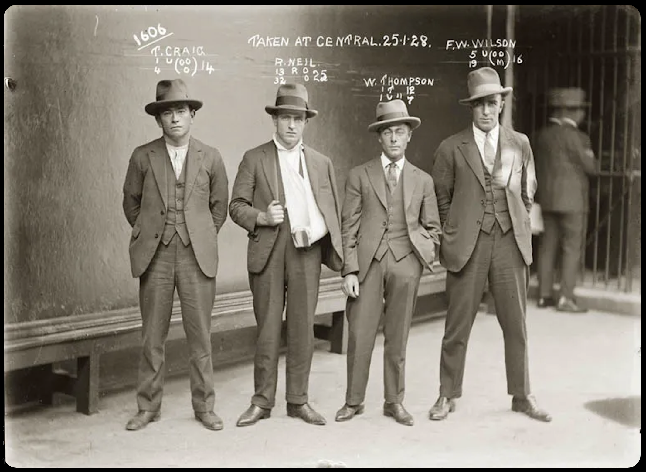 A black and white photograph of four young men dressed in early 20th-century suits and hats, standing in front of a wall with wooden paneling. Handwritten annotations above each man include names, dates, and other information.