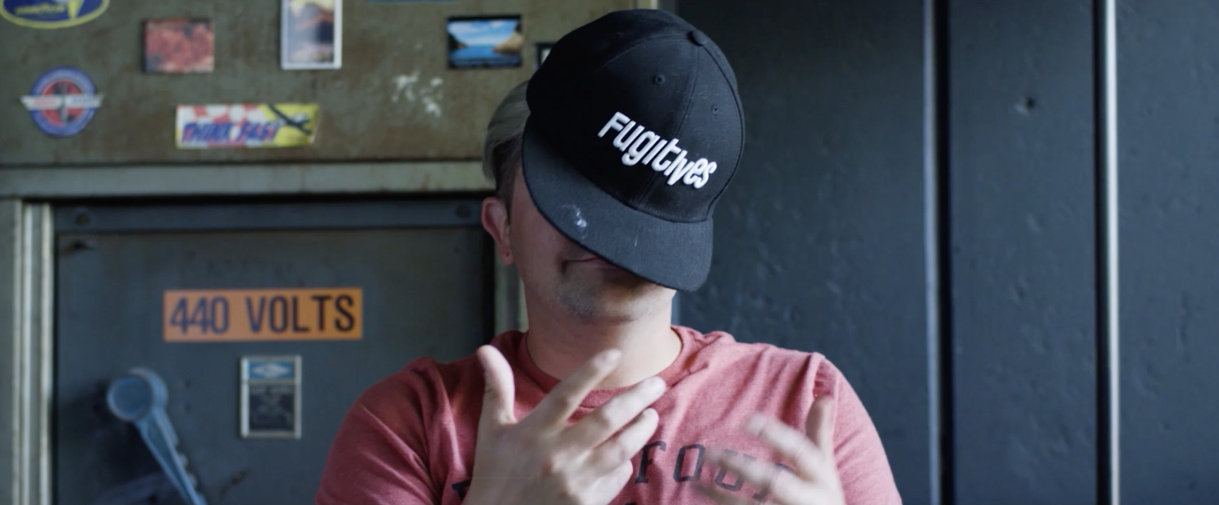 Person wearing a black cap with white writing that says "F*** Give" and a red T-shirt, with their face partially obscured by the cap, standing in front of a metal electrical box labeled "440 VOLTS".