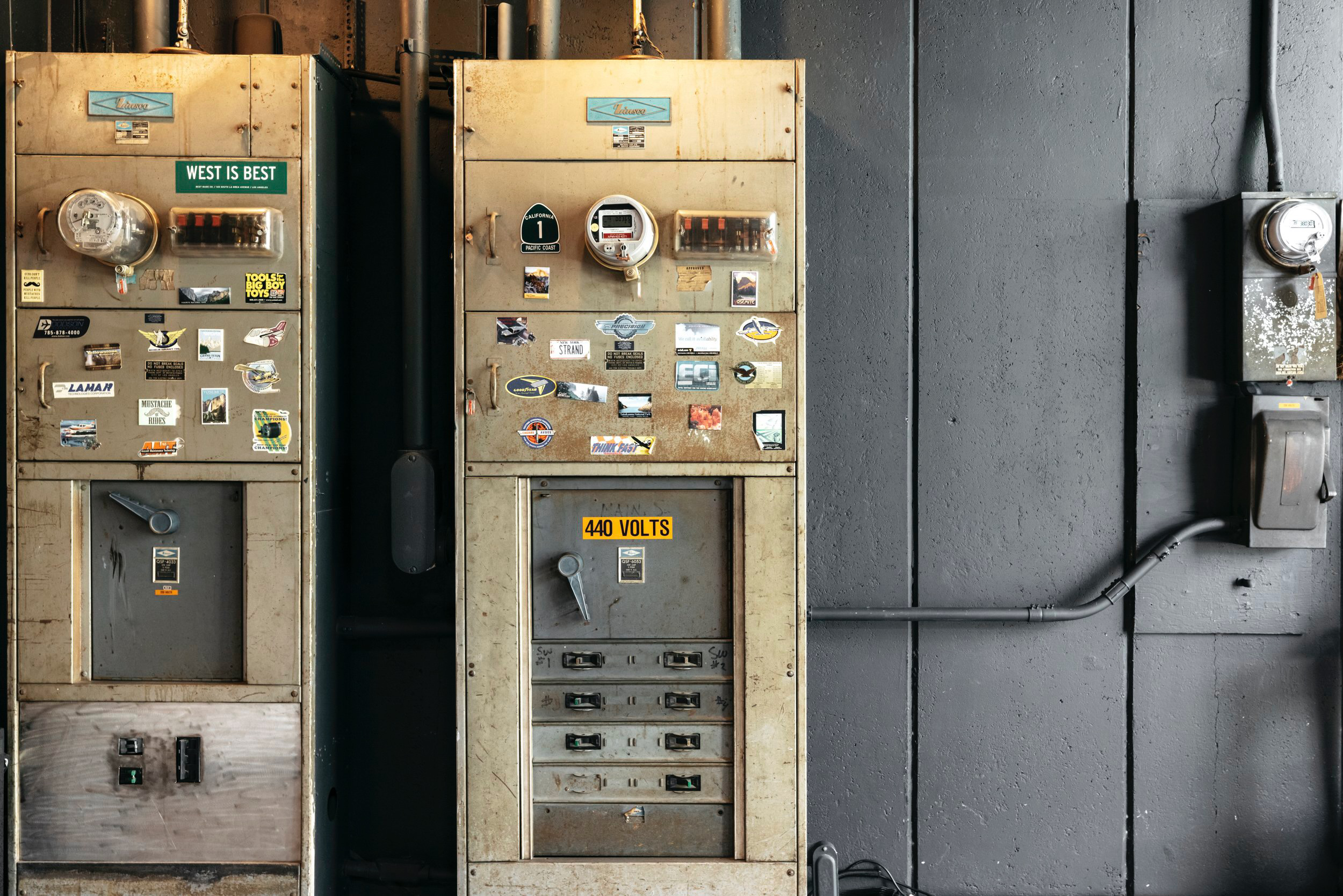 Two old utility meters with stickers on their surfaces are mounted on a dark wall. One meter has a sign that reads 'WEST IS BEST,' and the other displays '440 VOLTS.' To the right, a smaller electrical box is connected with a conduit pipe.