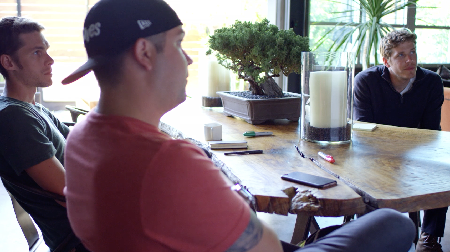 Three young men sit around a wooden table in a sunlit room, with plants and windows in the background, engaging in a discussion.