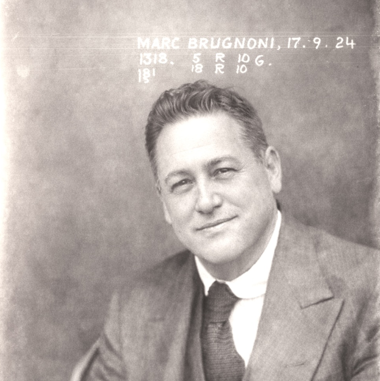 Black-and-white portrait of a man in a suit and tie, smiling, with writing above his head indicating his name, Marc Brugnoni, and some numbers and measurements.