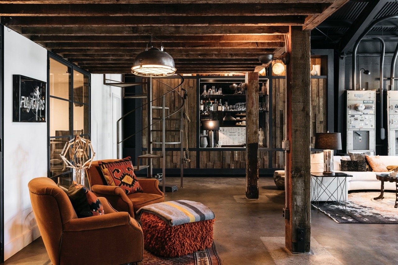A cozy, rustic living room with exposed wooden beams, a white sofa, orange armchairs, and a bar area in the background. Decor includes patterned cushions, a geometric sculpture, and warm lighting.