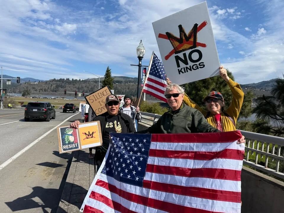 Group of protesters holding American flags and signs protesting against monarchy and promoting resistance, including a sign with a crossed-out crown and the words 'NO KINGS'.