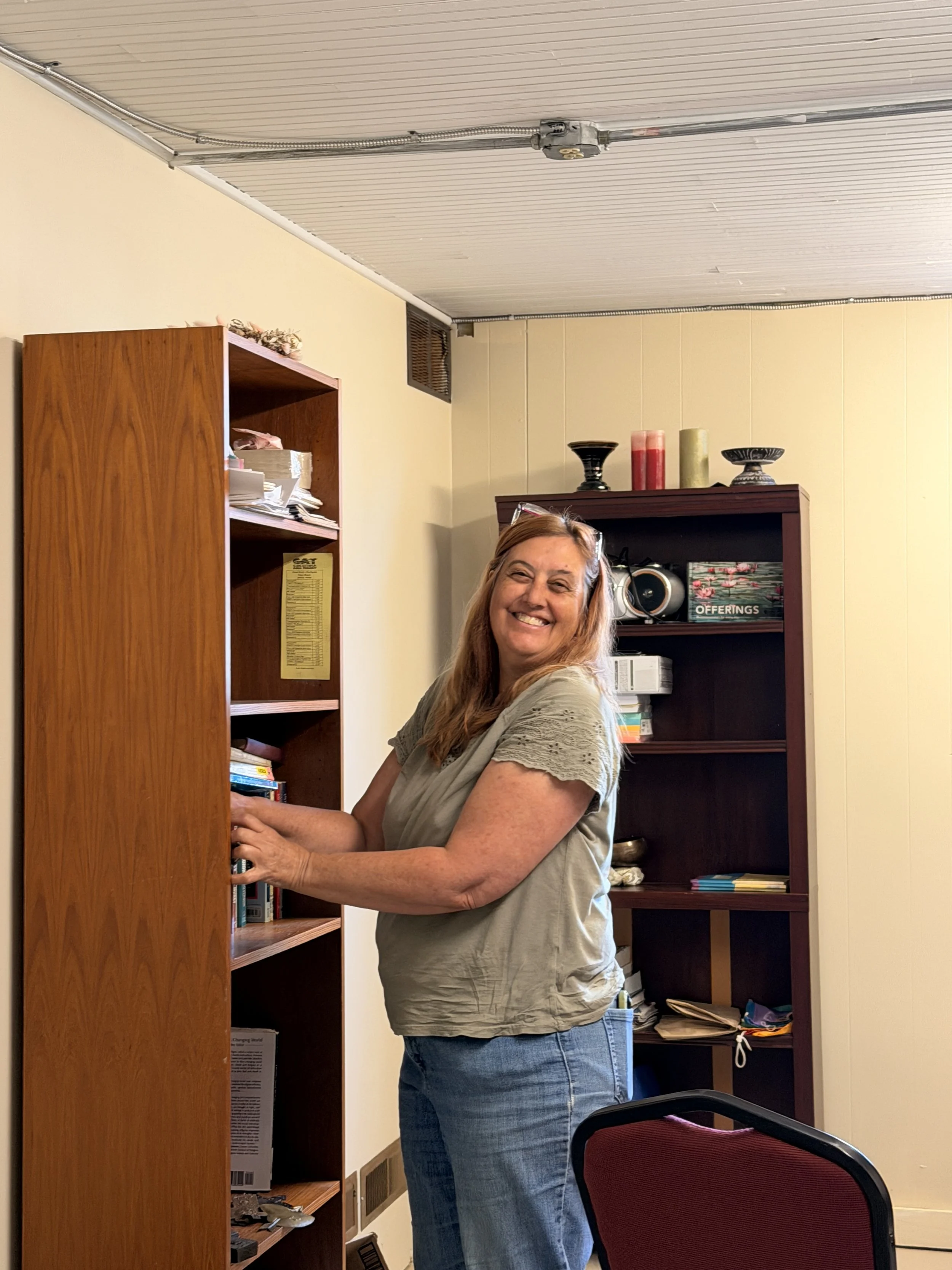 A woman with glasses on her head smiling as she stands by a wooden bookshelf filled with papers and books. She is wearing a light-colored top and jeans, and there are two bookshelves behind her with decorative candles and other items.