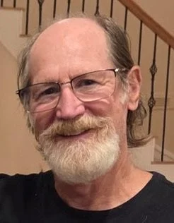 A middle-aged man with glasses, a beard, and mustache, smiling indoors with a staircase in the background.