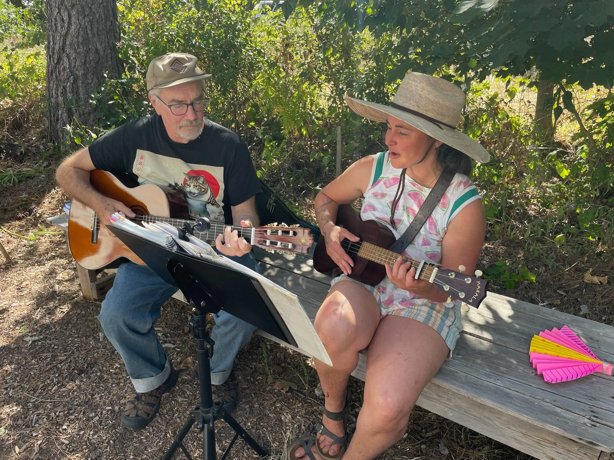 Two people sitting outdoors playing guitars, one man and one woman. The man has glasses and a hat, the woman has a wide-brimmed hat. They are sitting on a wooden bench with green foliage in the background. There are music sheets, pens, and colorful paper objects near them.