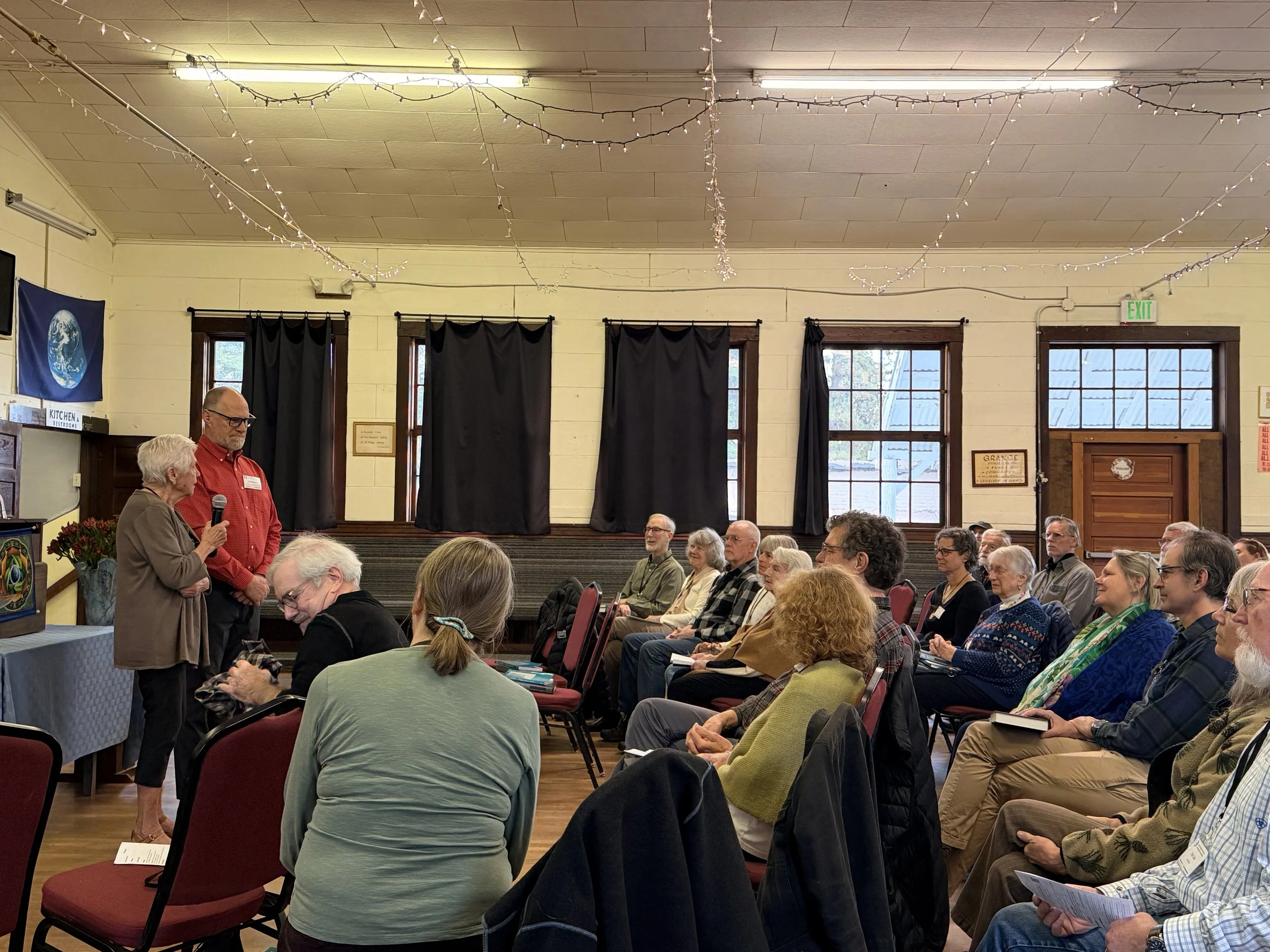 A room filled with an audience, primarily older adults, seated and listening to a woman speaking into a microphone. Two men stand at the front of the room, engaged in conversation. The room is decorated with string lights, black curtains, and a flag on the wall. Windows with dark curtains and an exit sign are visible.
