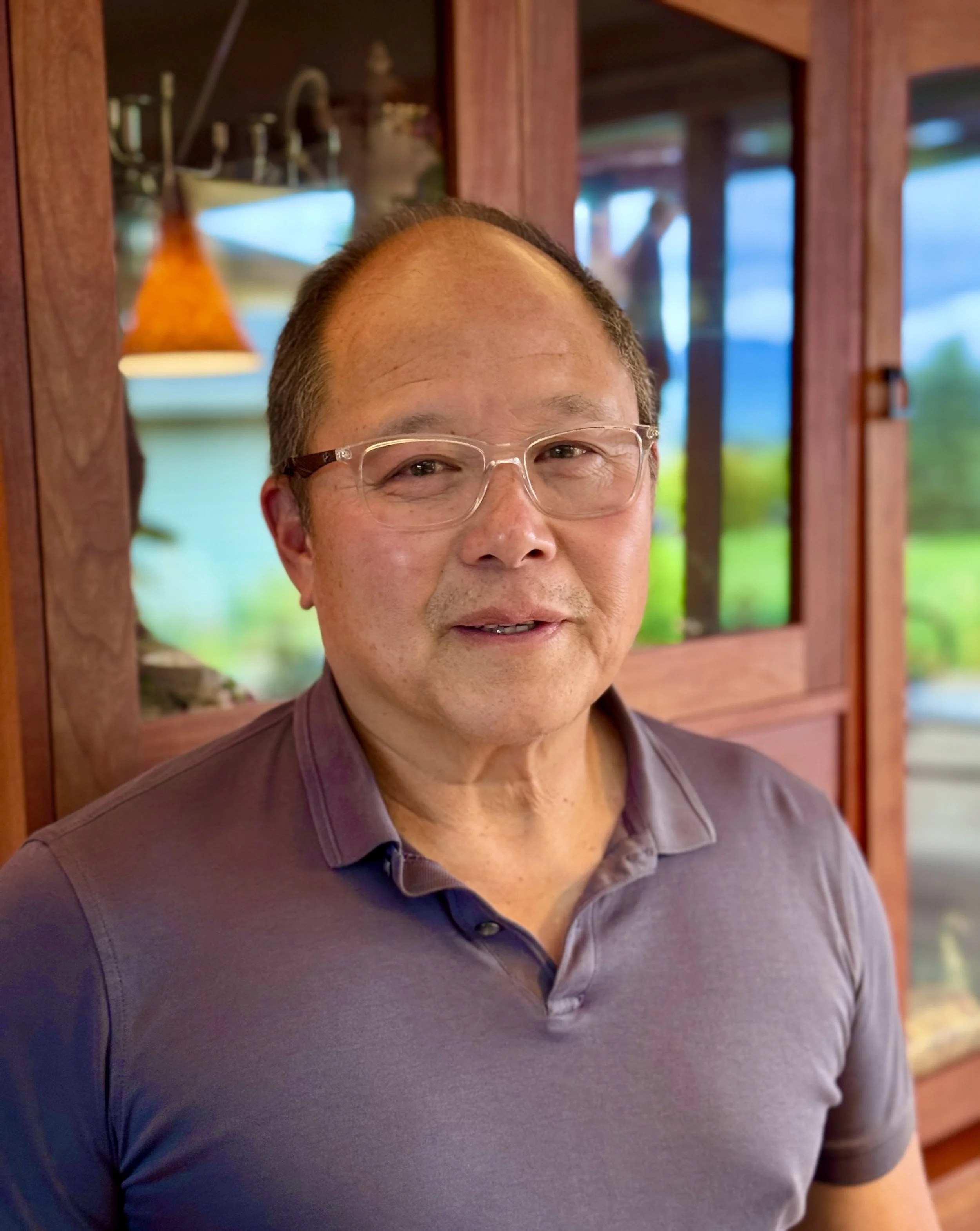 A close-up of a middle-aged man with glasses, smiling, standing in front of a wooden and glass cabinet with a blurred background of outdoor scenery.