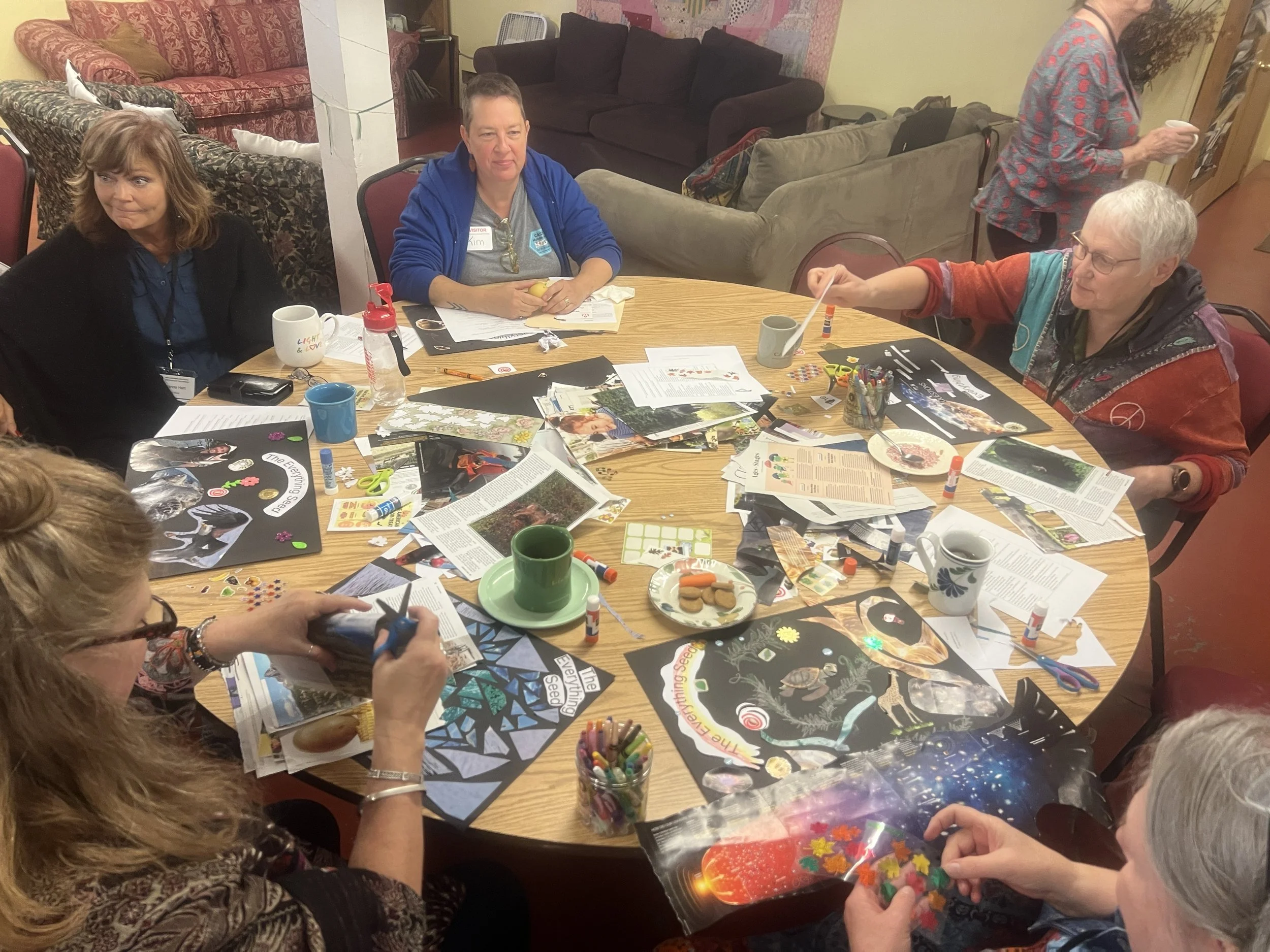 A group of people are sitting around a large wooden table engaged in arts and crafts activities. The table is cluttered with paper, magazines, scissors, glue, stickers, and craft supplies. There are three women visible, one with glasses, and a woman with a blue jacket and a name tag labeled 'Kim.' In the background, there are couches, a fan, and a person holding a cup, with various paper and fabric on the couches and walls.